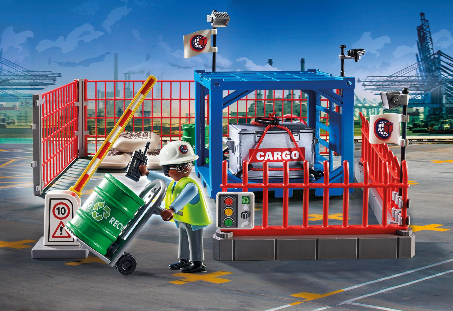 Eine Spielzeugszene zeigt einen Arbeiter mit einer Recyclingtonne, einem Frachtcontainer und Barrieren unter blauem Himmel. Es gibt Flaggen und Schilder.