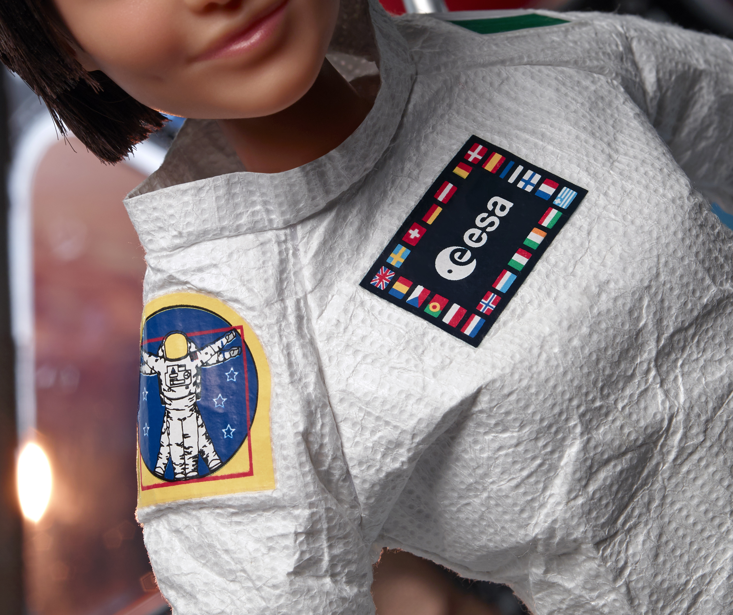 Muñeca con traje de astronauta. Tejido blanco texturizado. Parches con banderas y logotipos. Pelo corto castaño. Sonrisa.