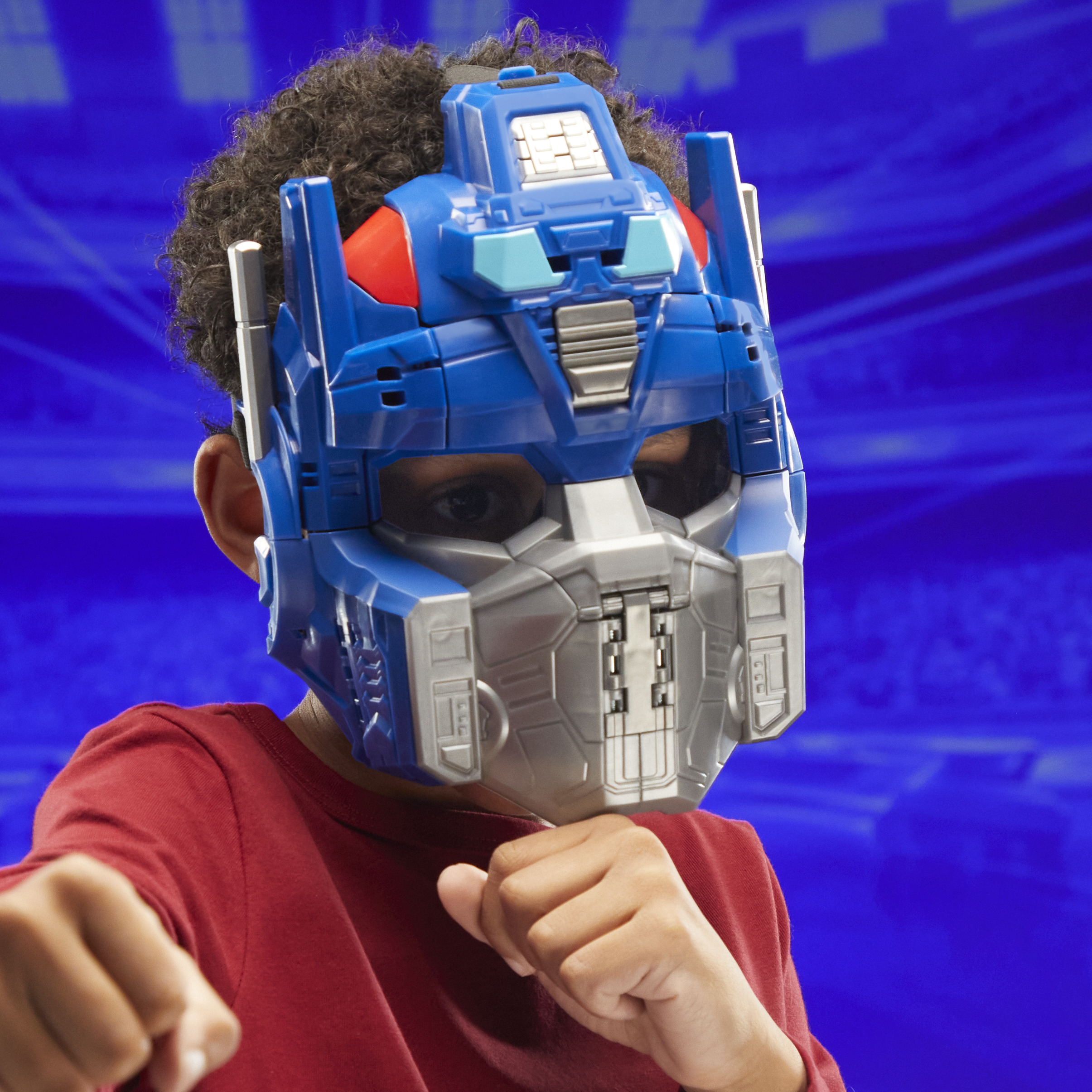 Un niño con una máscara de Optimus Prime y una camisa roja, haciendo un puño. Fondo azul.