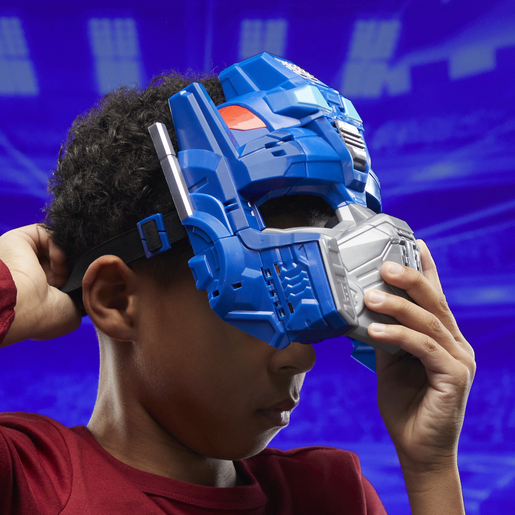 Un niño lleva una máscara de Transformer azul y plateada. El niño sostiene la máscara contra un fondo azul.
