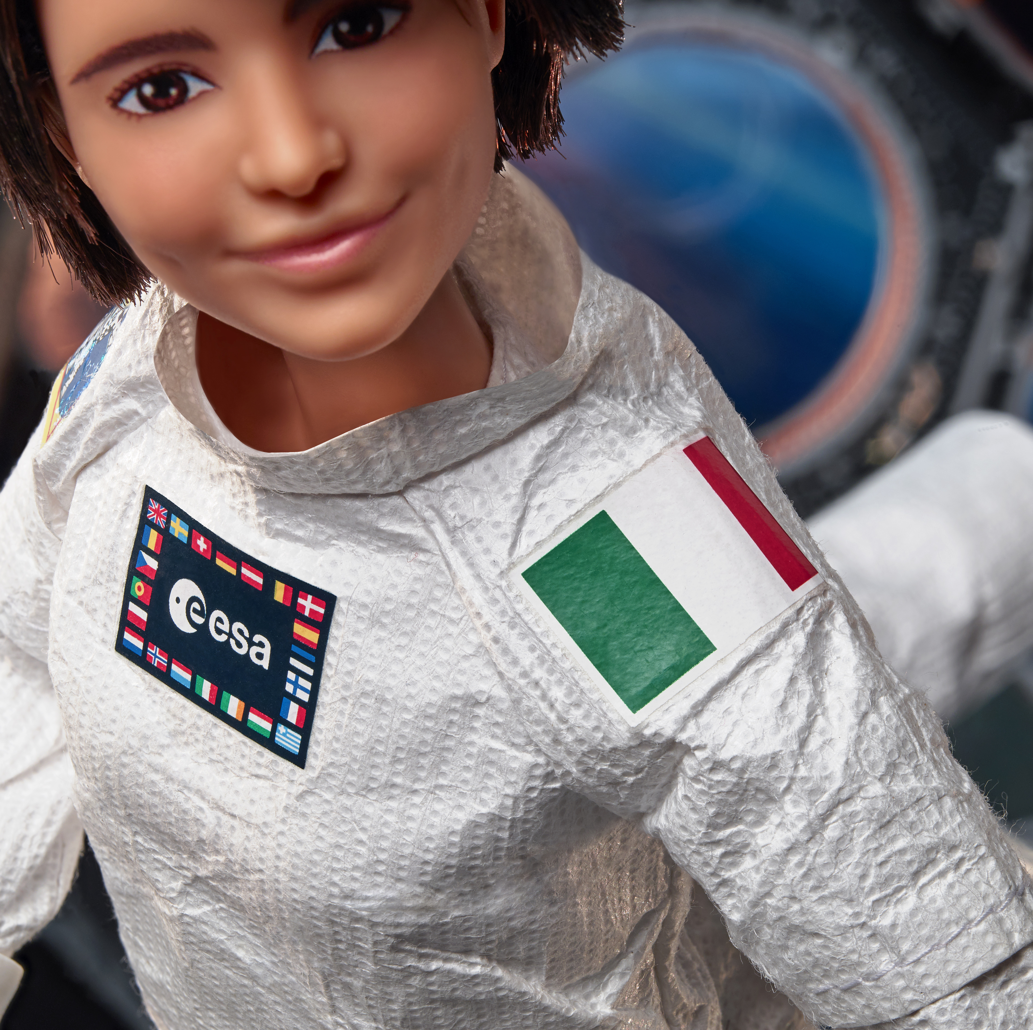 Una muñeca con un traje espacial blanco con parches de la ESA y la bandera italiana. La muñeca tiene cabello y ojos marrones, sonriendo.