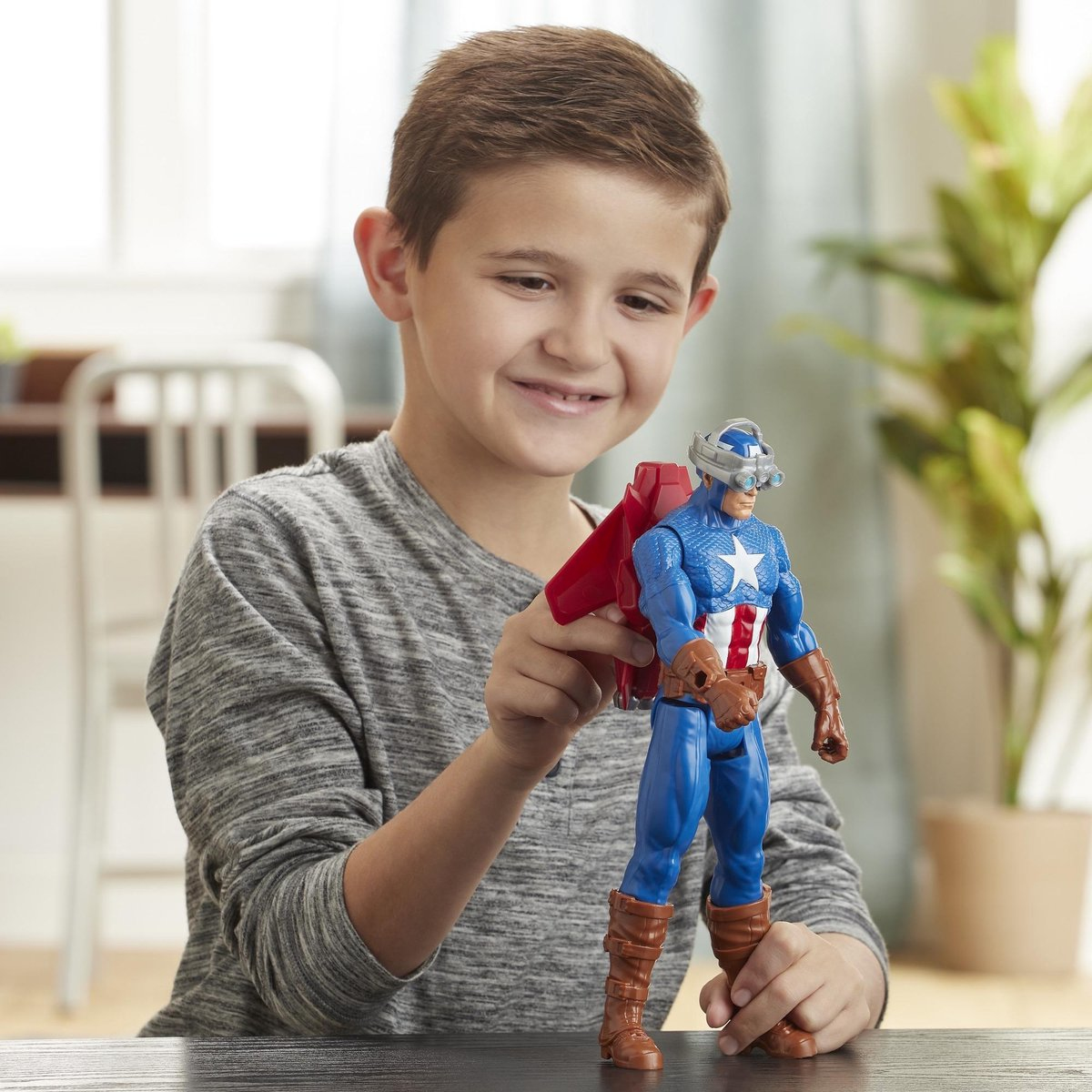 Un ragazzo tiene in mano un personaggio di Captain America con un'ala rossa. Il ragazzo sorride. Una sedia, un tavolo e una pianta sono sullo sfondo.
