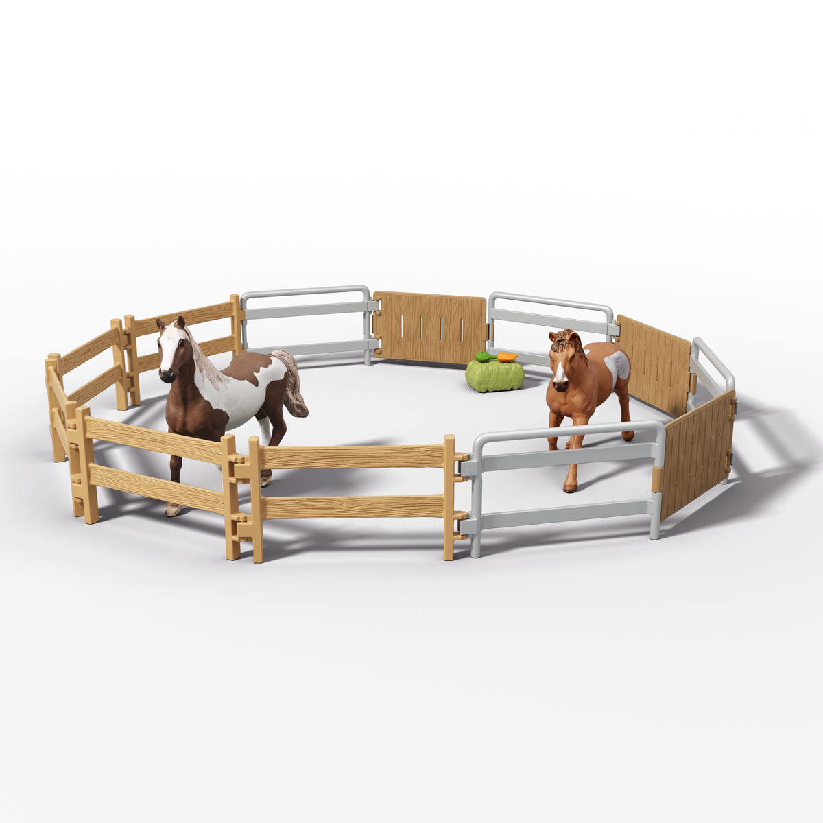 Dos caballos de juguete en un corral circular. Uno es marrón y blanco, el otro marrón. Hay una paca de heno y una zanahoria.