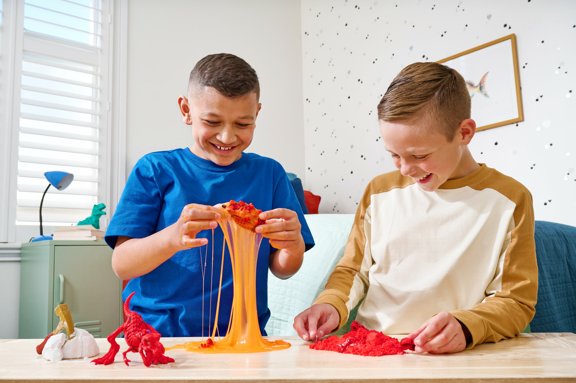 Dos niños juegan con slime y dinosaurios de juguete en una mesa de madera. Uno estira slime naranja, el otro trabaja con rojo.