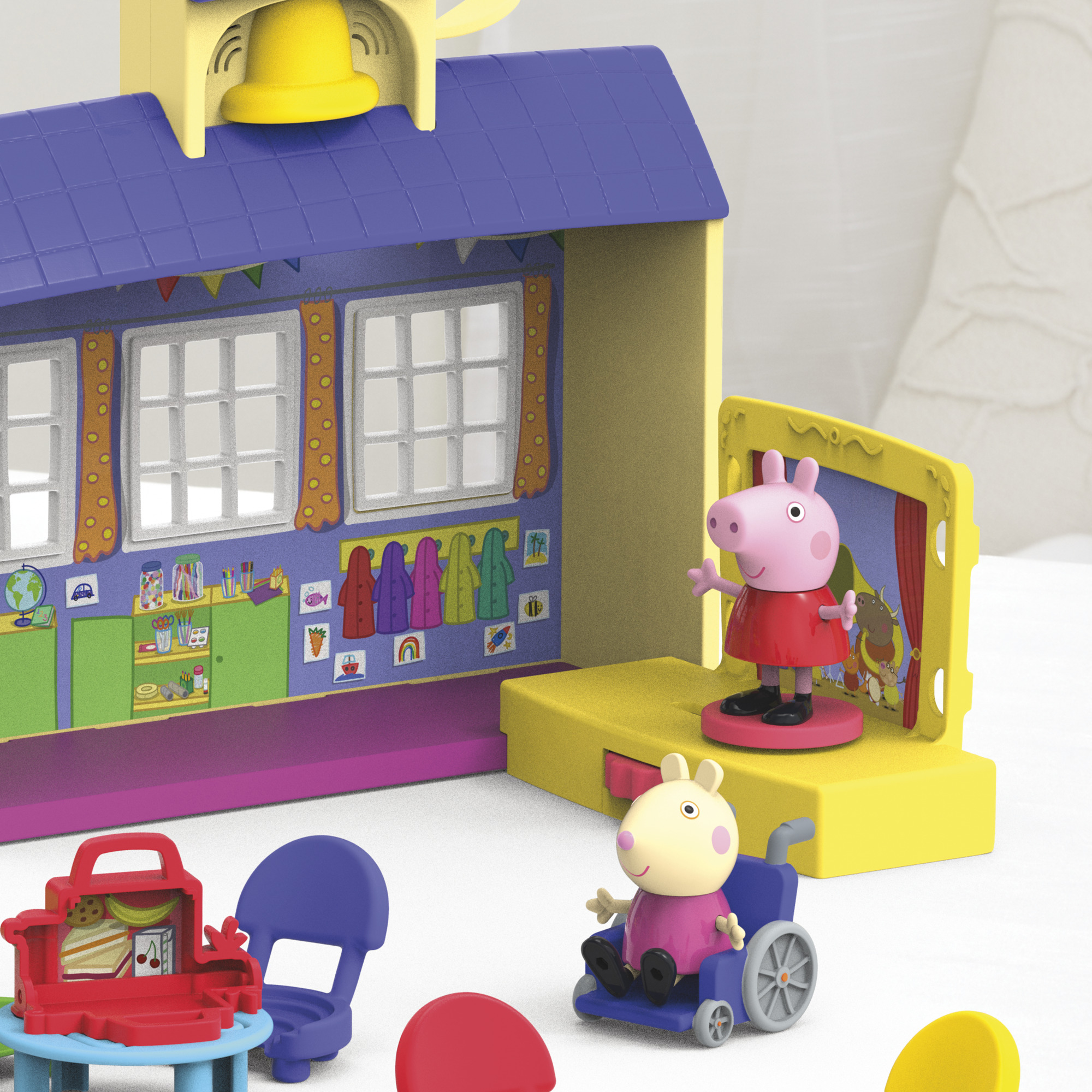 Ein Peppa Wutz Spielset. Es zeigt ein Schulgebäude, eine Bühne, Peppa Wutz Figuren und einen Rollstuhl. Eine Brotdose und Stühle sind im Vordergrund.