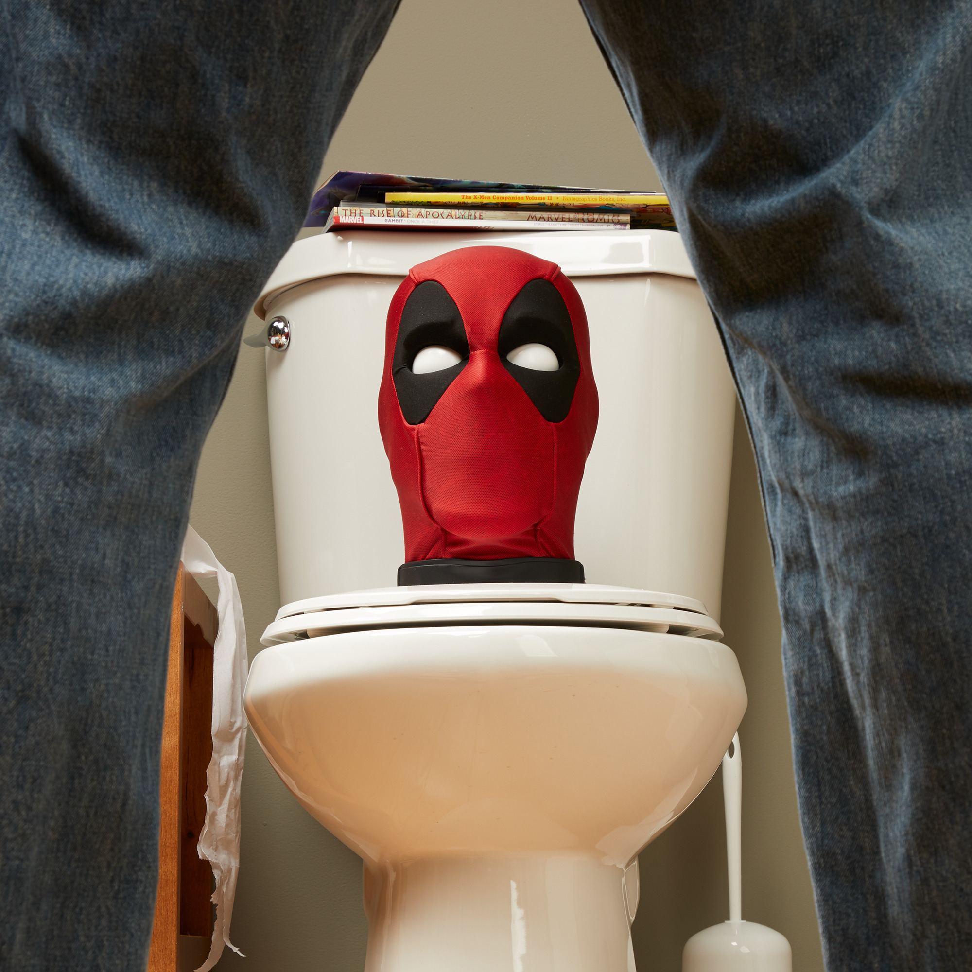 Máscara de Deadpool en un asiento de inodoro, rodeada de jeans. Cómics en el tanque. Papel higiénico y un desatascador son visibles.