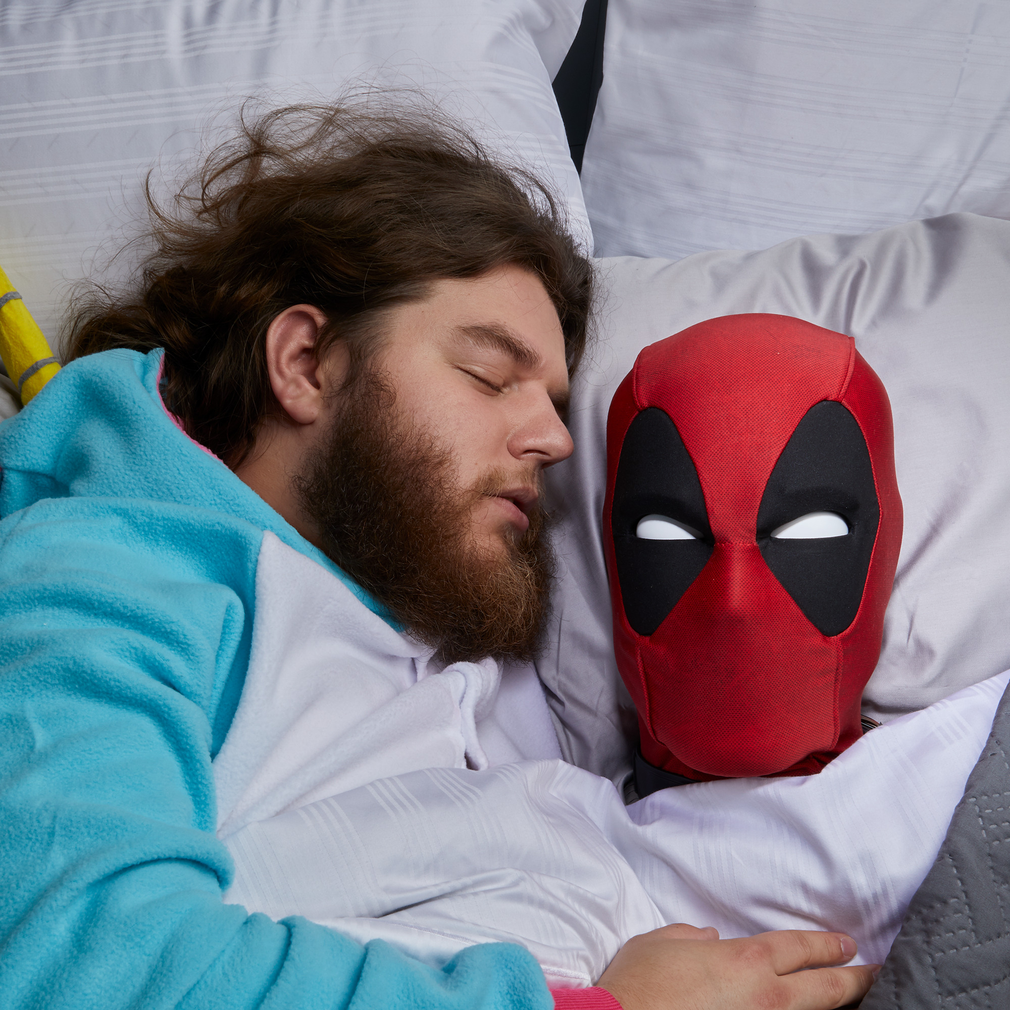Un hombre con un mono de unicornio duerme junto a una máscara de Deadpool en una cama con ropa de cama blanca y gris.