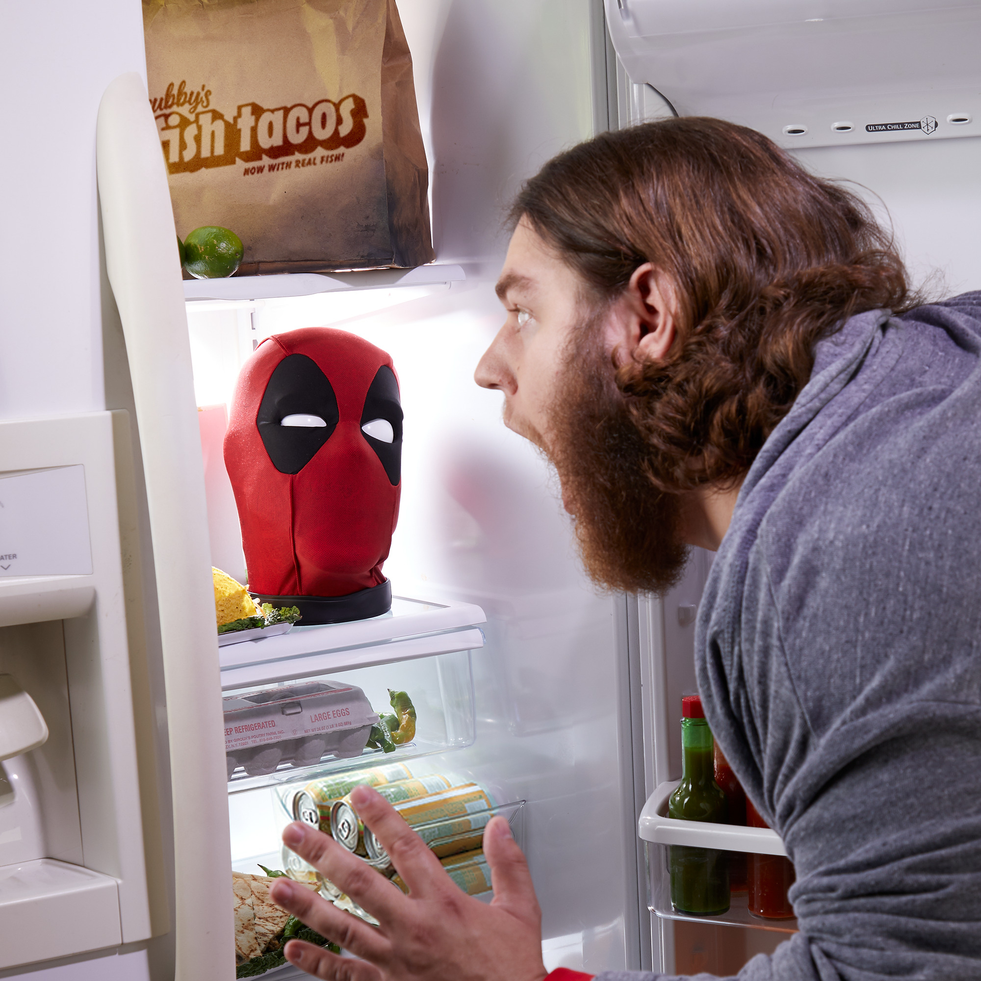 Un hombre mira sorprendido una máscara de Deadpool en una nevera. La nevera contiene alimentos como huevos y tacos de pescado.