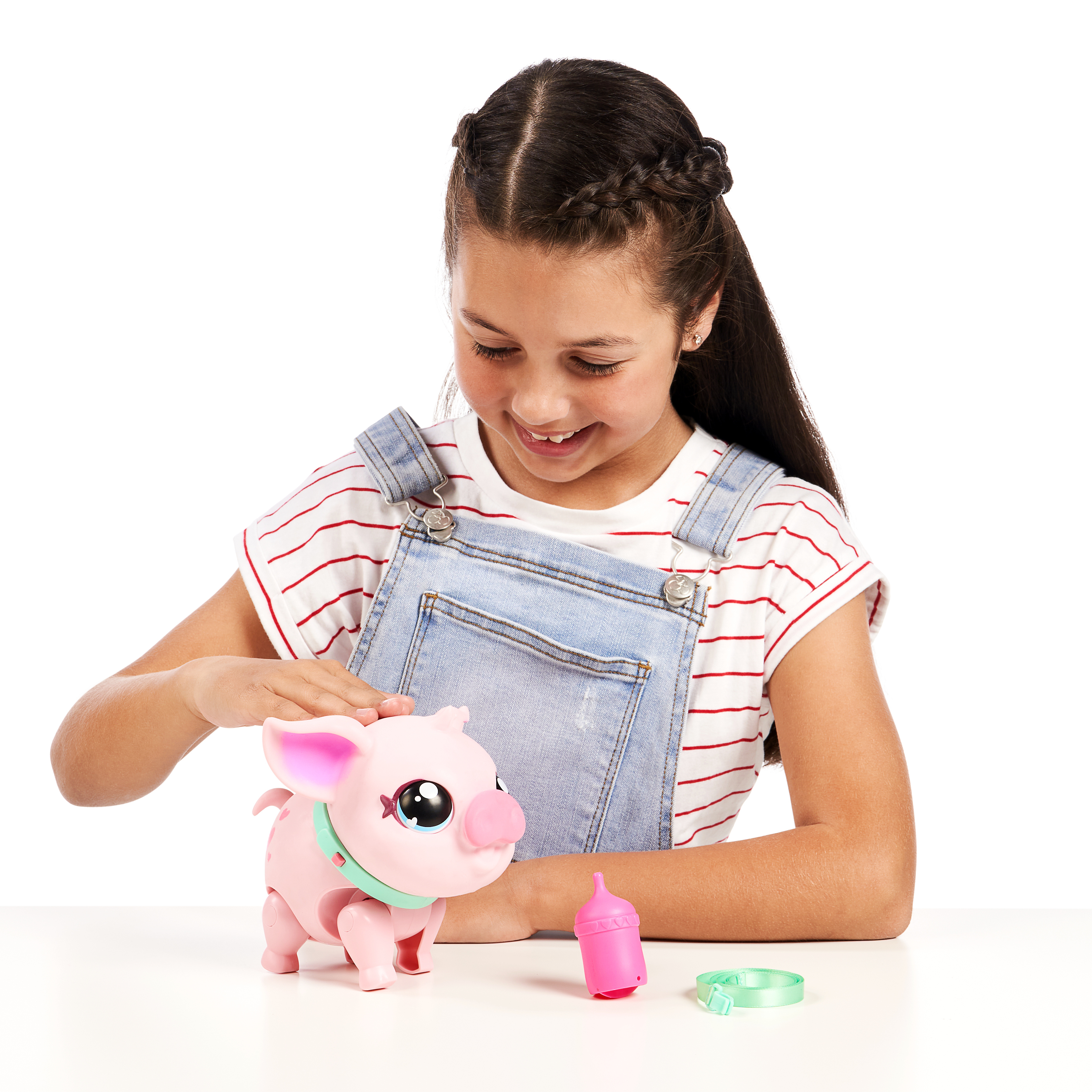 Ein Mädchen lächelt ein rosa Schweinespielzeug an. Auf dem Tisch liegen eine Flasche und ein Halsband. Das Mädchen trägt eine Latzhose und ein gestreiftes Hemd.