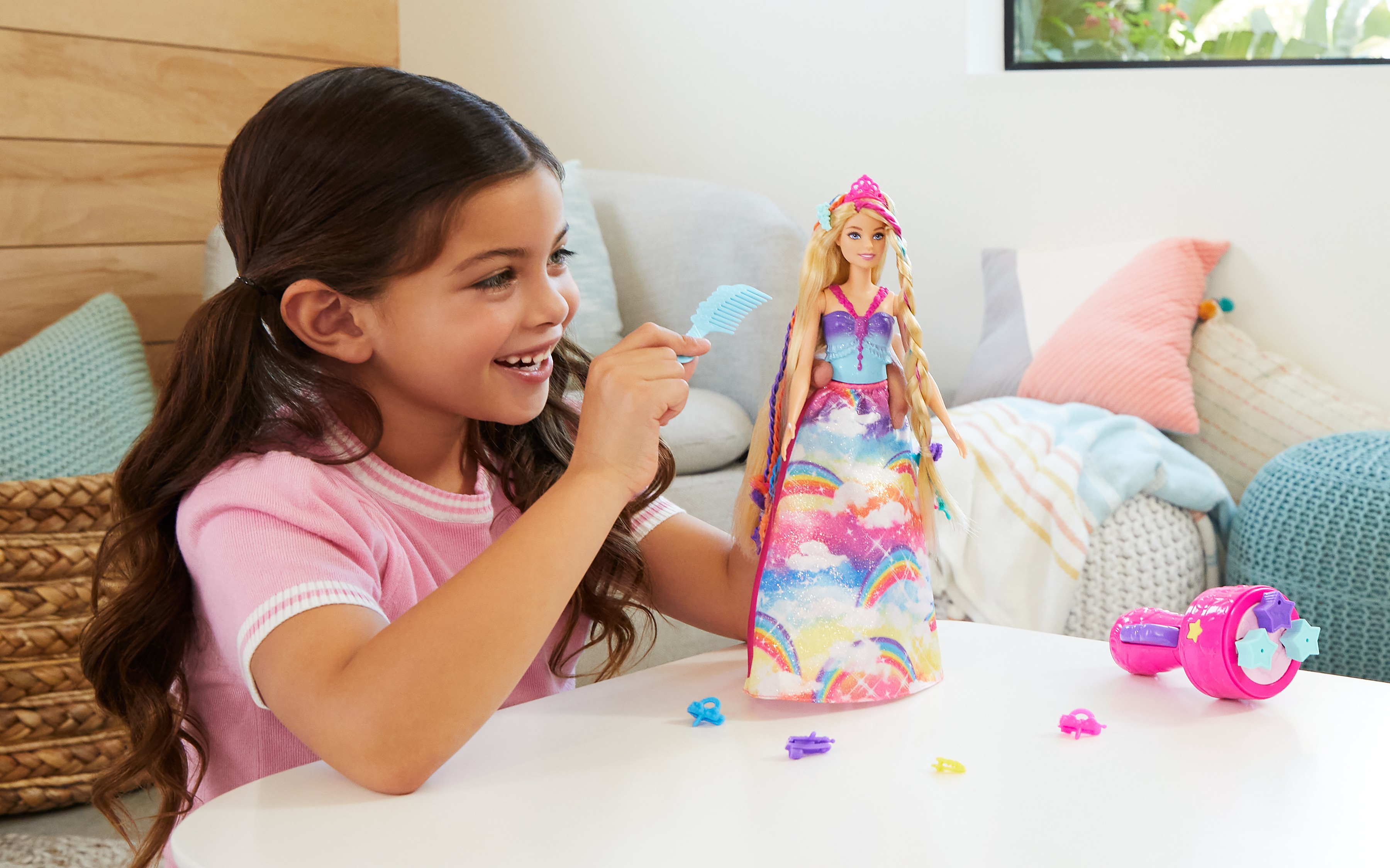 Una bambina pettina i capelli di una bambola. La bambola ha un vestito arcobaleno. Giocattoli e cuscini sono sul tavolo e sullo sfondo.