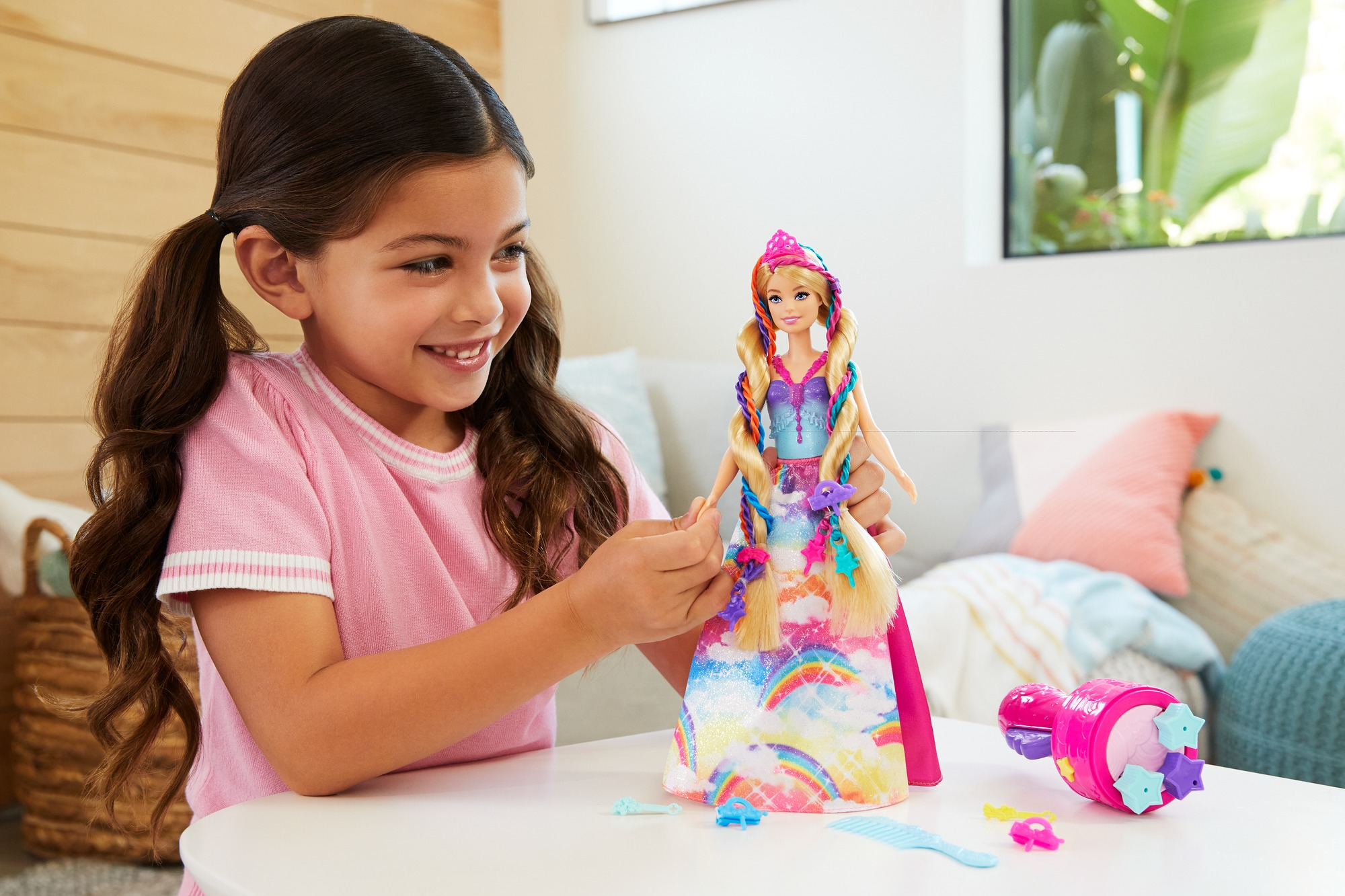 Una bambina sorride mentre gioca con una bambola Barbie. La bambola ha lunghi capelli biondi. Ci sono accessori sul tavolo.