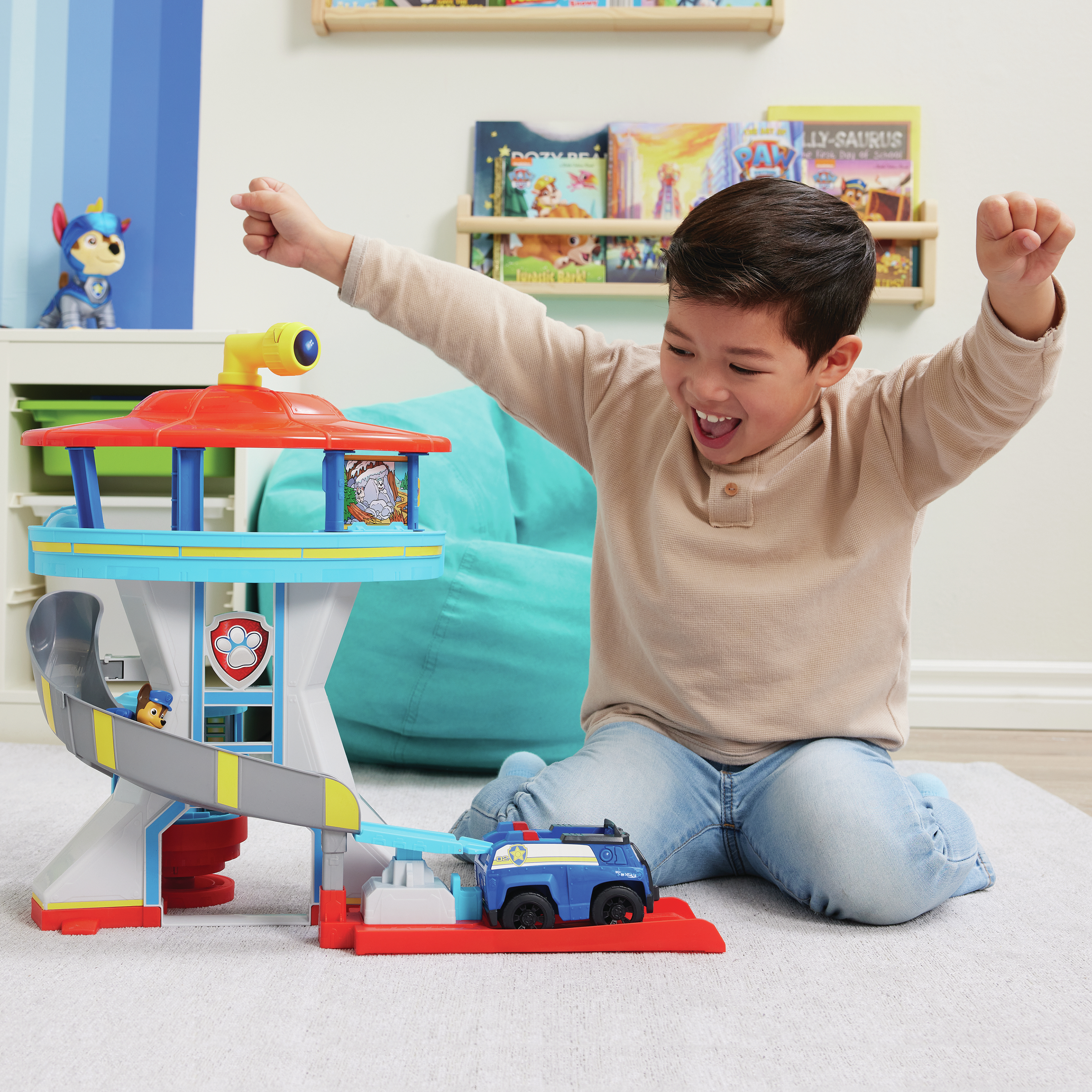 Un ragazzo esulta accanto a un giocattolo di Paw Patrol. Il giocattolo ha uno scivolo e un'auto. Ci sono librerie sullo sfondo.