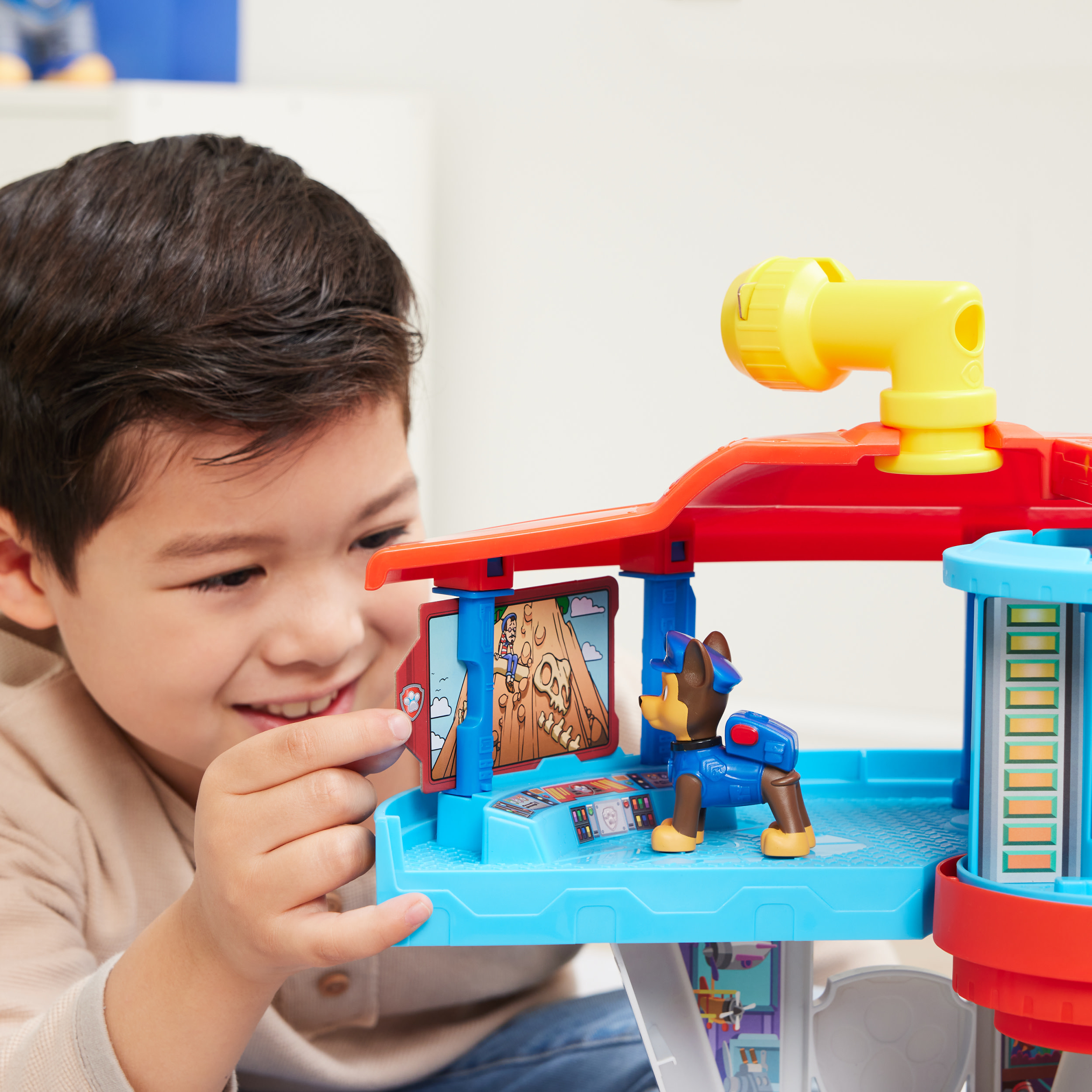 Un ragazzo sorride, guardando un playset giocattolo con un personaggio di Chase. Il set ha componenti rossi e blu.