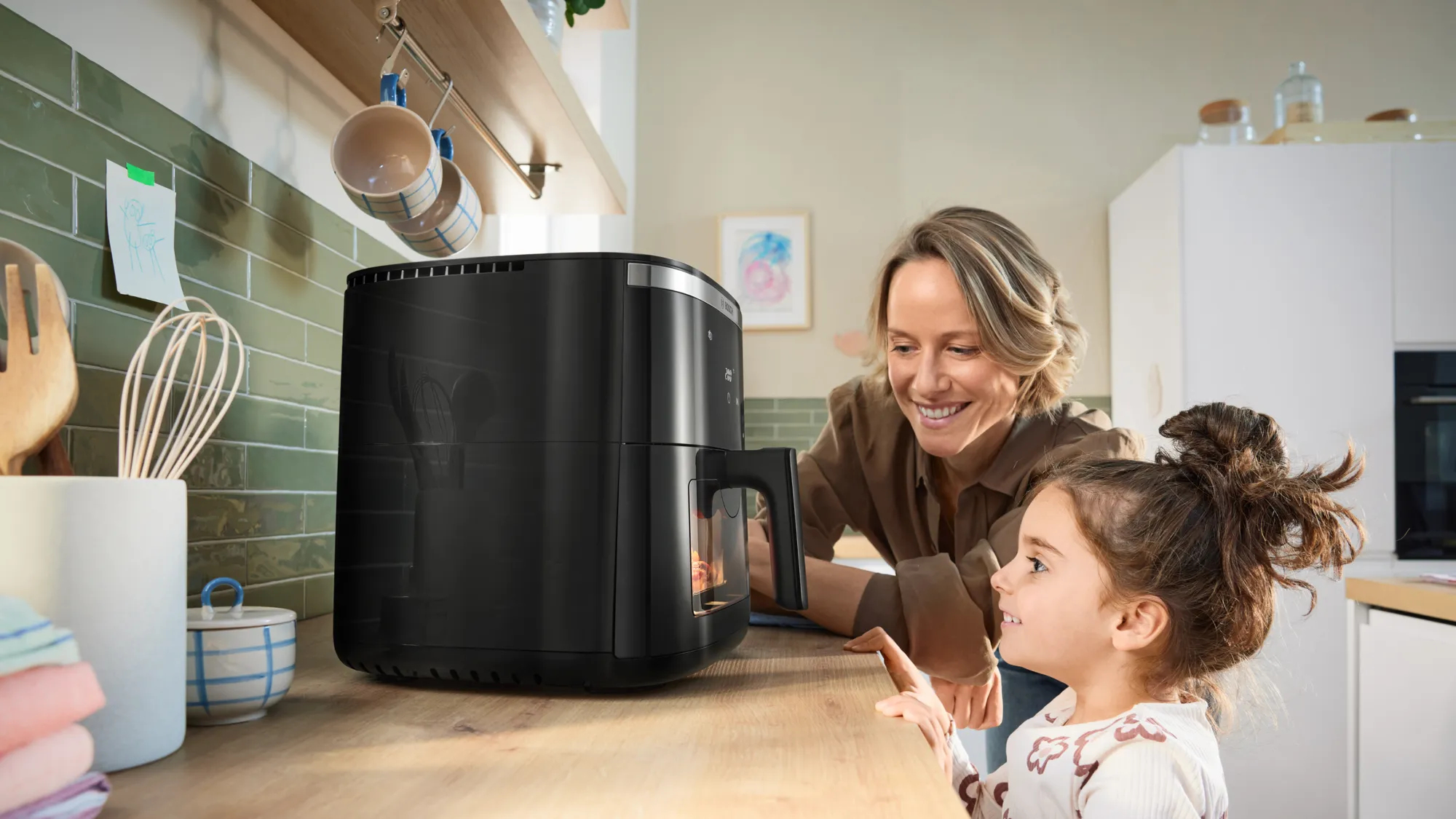 Kobieta i dziewczynka patrzą na czarną frytkownicę powietrzną na drewnianym blacie w kuchni. Kobieta się uśmiecha.