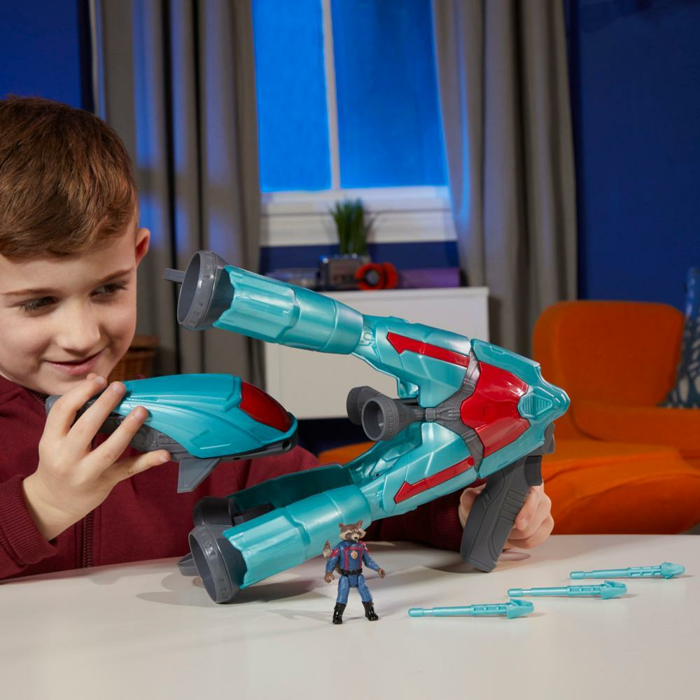 Un niño sostiene una pistola de juguete con diseño rojo y azul. Una figura de Rocket está cerca. Una silla naranja brillante está atrás.