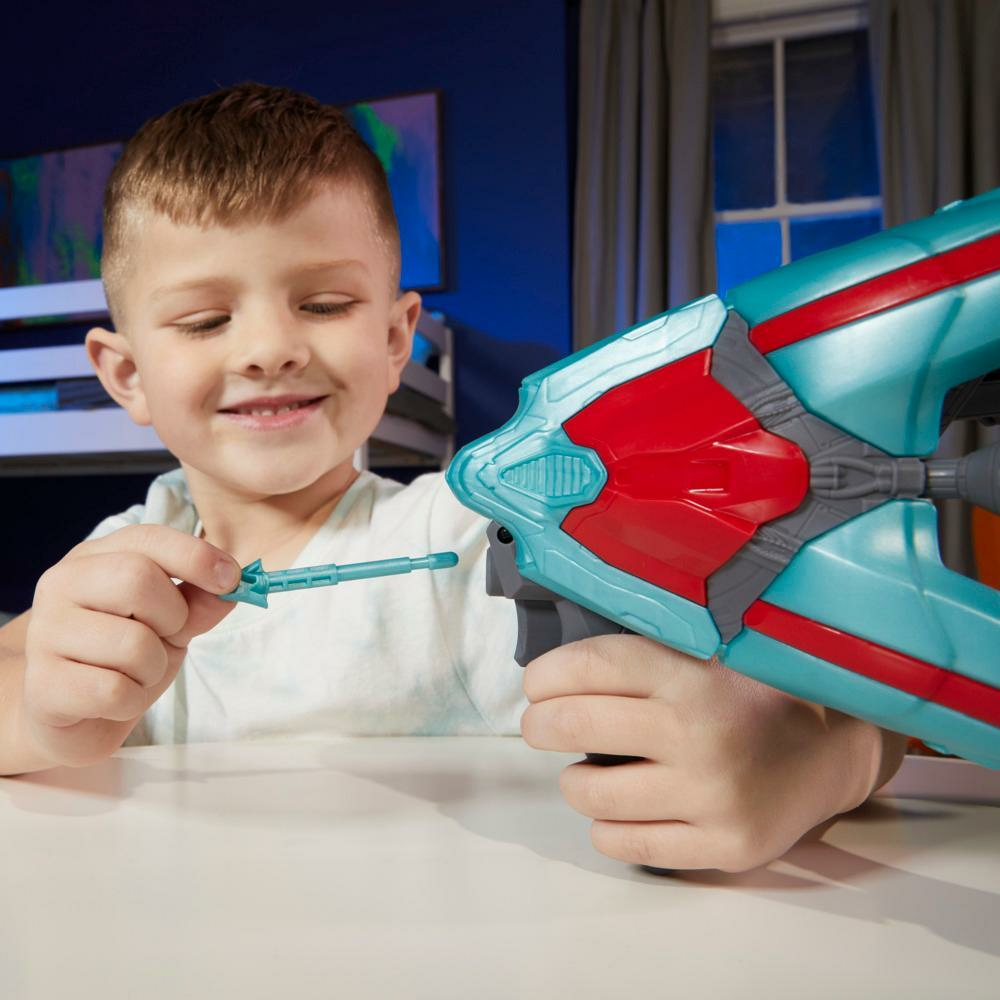 Un niño sonriente sostiene una pistola de juguete. La pistola es azul y roja. Sostiene un pequeño dardo. Hay una mesa blanca frente a él.