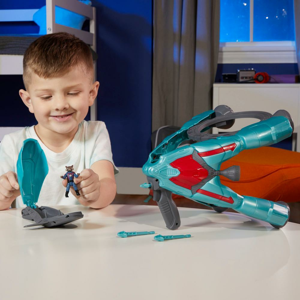 Un niño sonríe, sosteniendo una figura de Rocket Raccoon. Una pistola de juguete y un contenedor están sobre la mesa.
