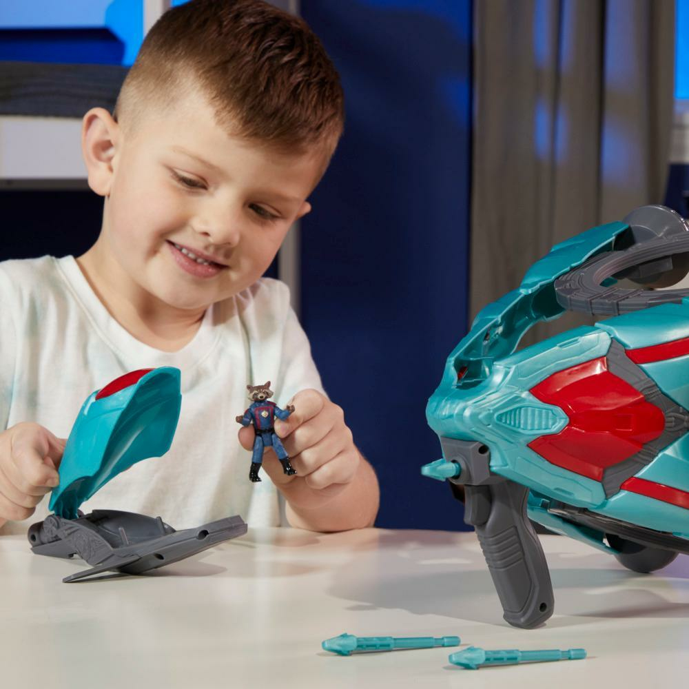 Un niño sonríe, sosteniendo una figura de juguete de Rocket. Está sentado en una mesa con una pistola de juguete y otras piezas de juguete.