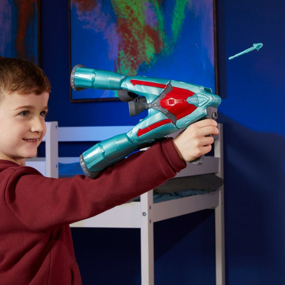 Un niño sostiene una pistola de juguete, apuntando. La pistola es azul y roja. Una flecha está en el aire. Se ve una litera y una pared azul.