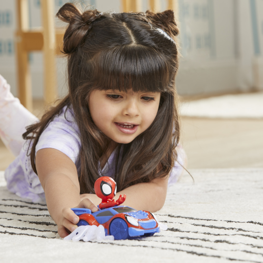 Dziewczynka bawi się samochodzikiem Spider-Mana. Samochód jest niebiesko-czerwony. Dziewczynka uśmiecha się i patrzy na samochód.