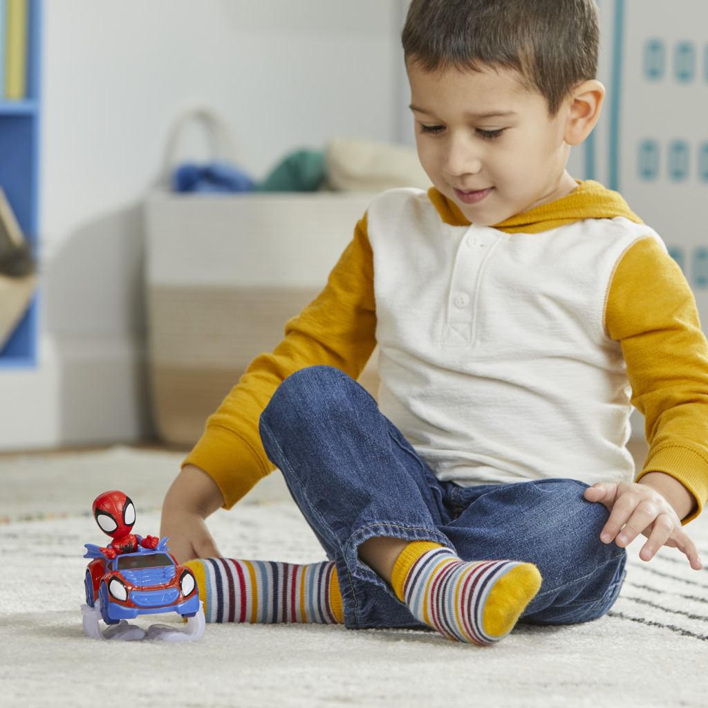 Chłopiec bawi się samochodzikiem Spider-Mana. Ma na sobie żółto-białą koszulę, dżinsy i paski. W tle niebieska półka.