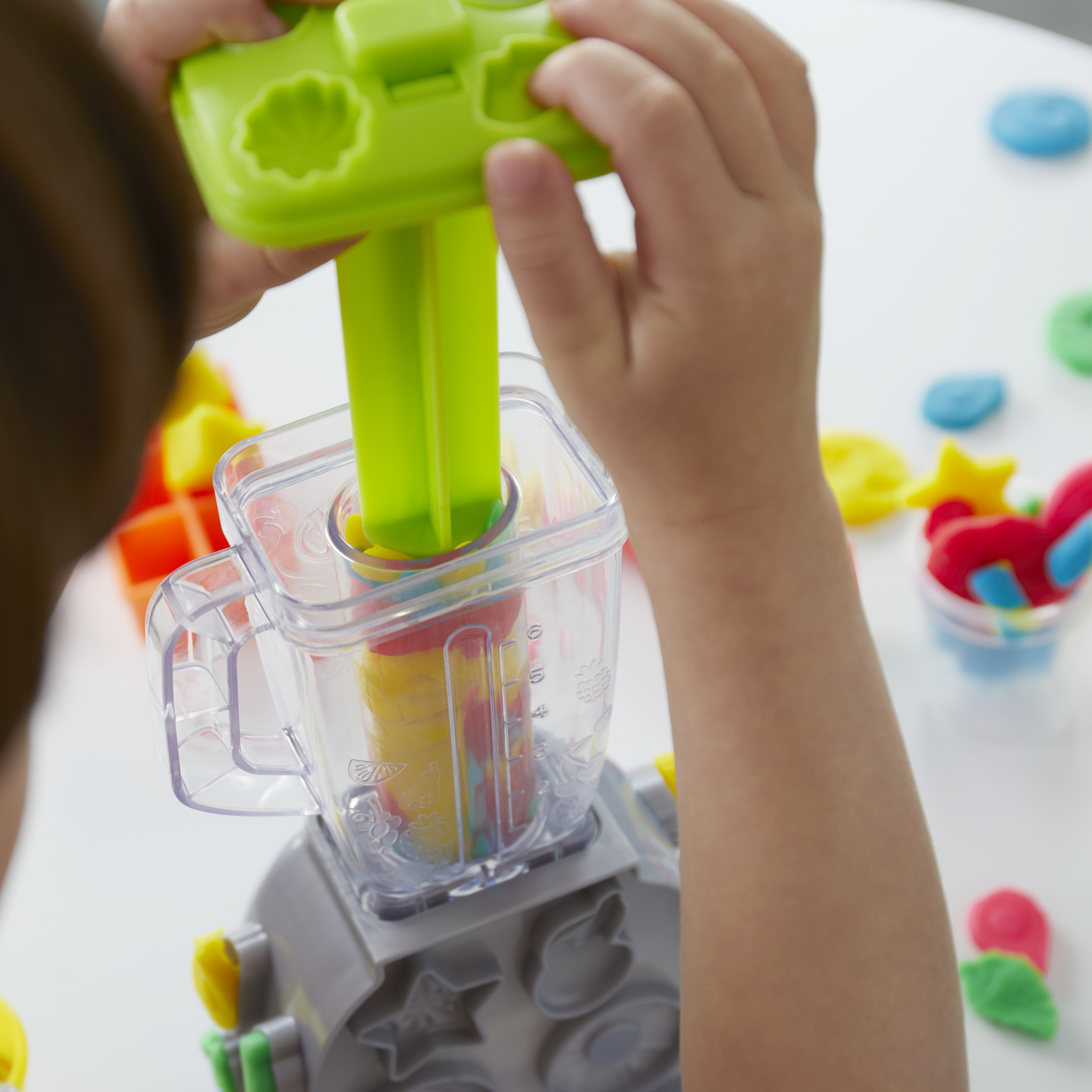 Un niño usa una herramienta verde de plastilina para presionar plastilina colorida en un juguete con forma de licuadora sobre una superficie blanca.
