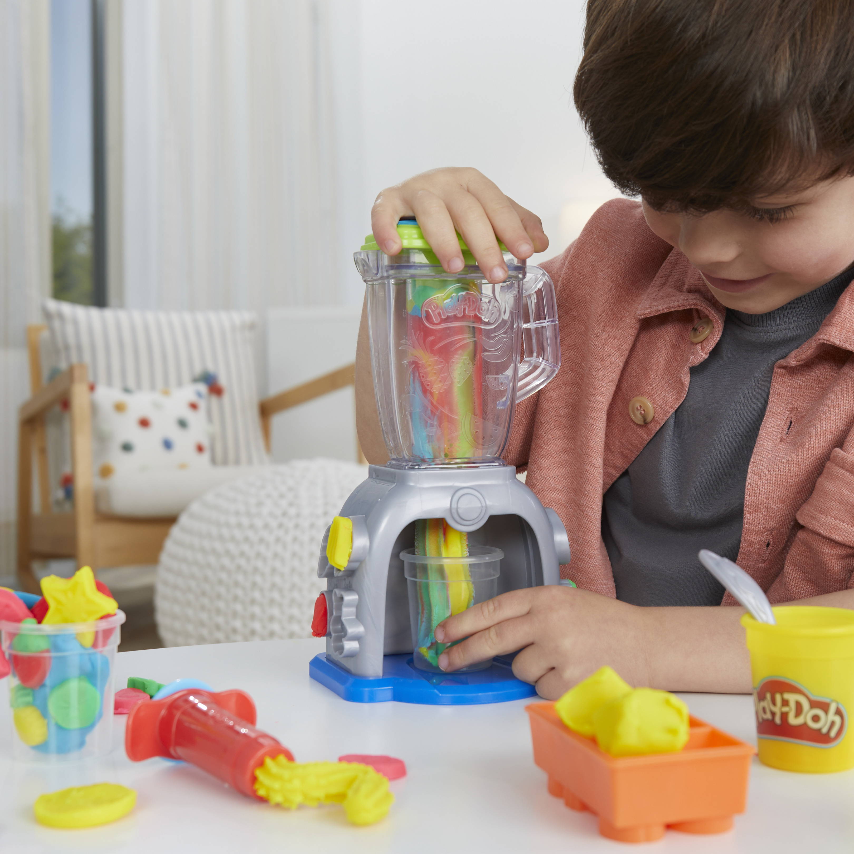 Un niño juega con Play-Doh. Usa una batidora de juguete con Play-Doh de colores. Hay otras herramientas y colores de Play-Doh en la mesa.