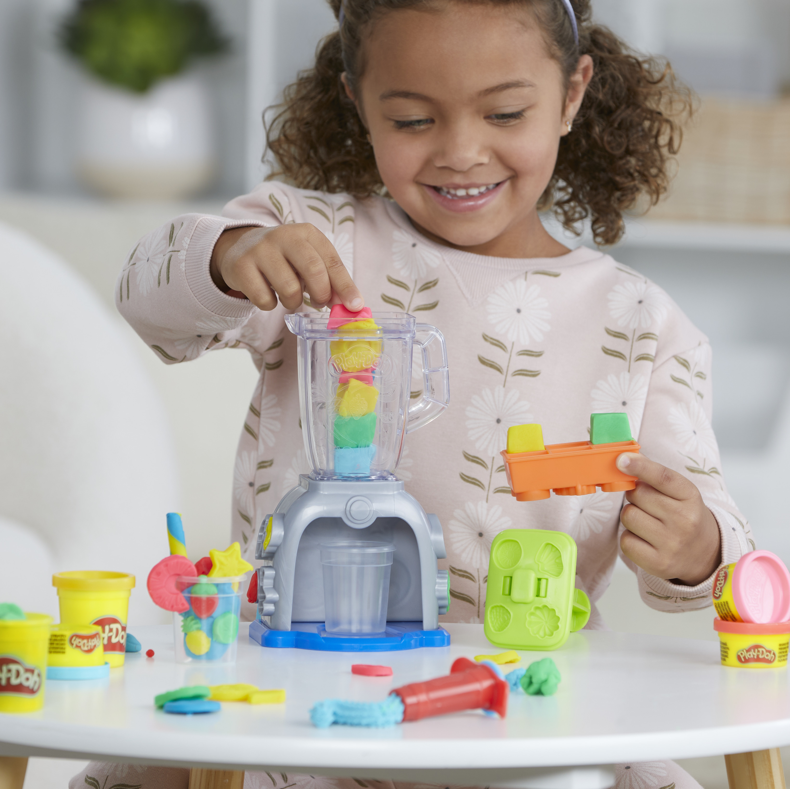Una niña juega con juguetes Play-Doh. Está poniendo un bloque rosa y amarillo en una licuadora de juguete, sonriendo.