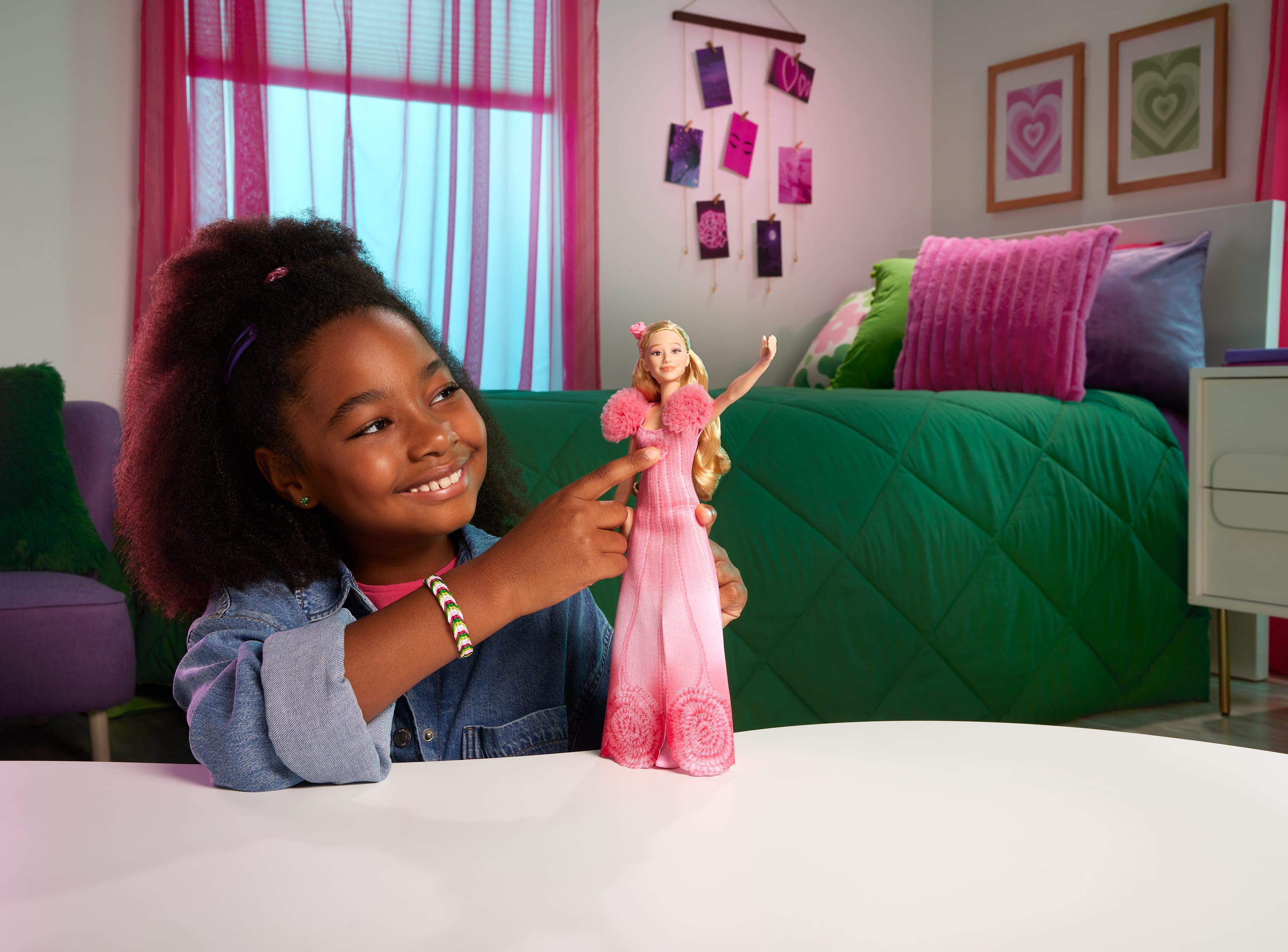 Ein Mädchen zeigt auf eine Barbiepuppe in einem rosa Kleid. Im Hintergrund sind ein Bett, Bilder und ein Fenster.