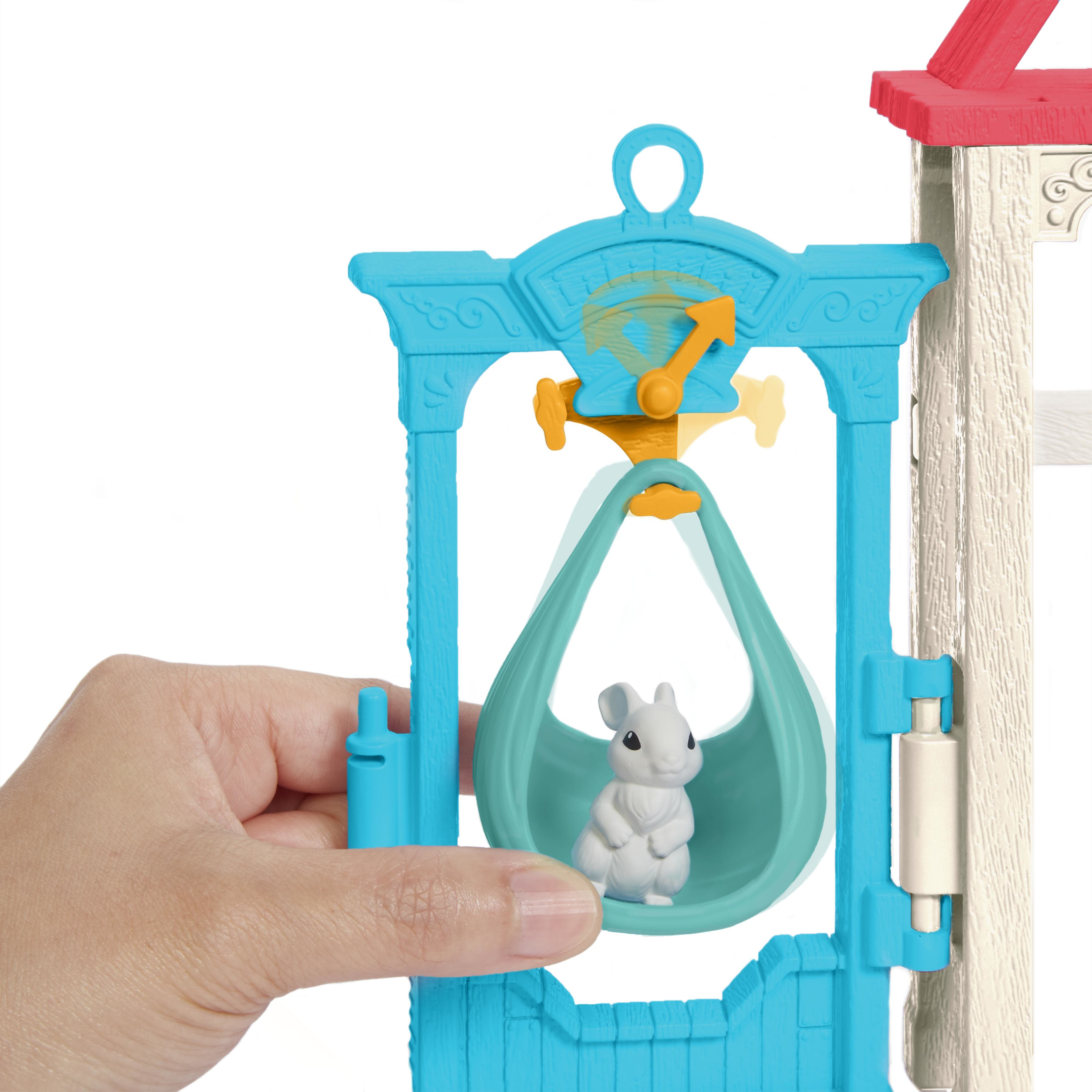 Ręka trzyma zabawkę. Mała biała mysz siedzi w turkusowej kołysce, w niebieskiej konstrukcji. Powyżej znajduje się zegar.