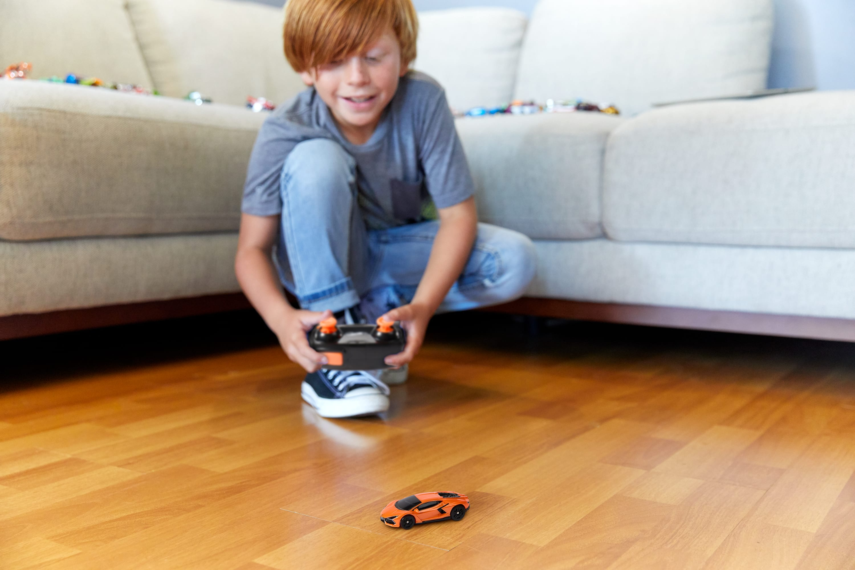 Chłopiec bawi się zabawką. Kuca, trzyma pilota. Pomarańczowy samochód stoi na drewnianej podłodze.