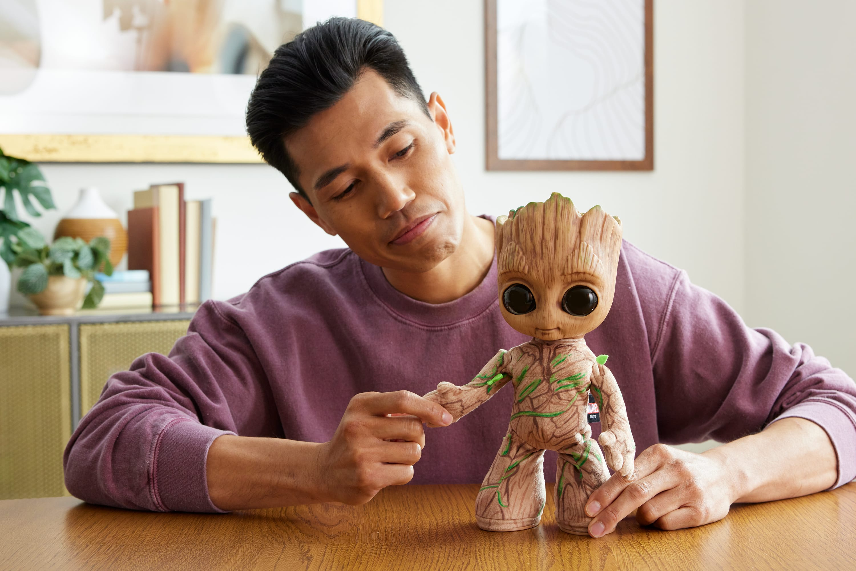 Mężczyzna patrzy na zabawkę Baby Groot. Ma na sobie fioletowy sweter i siedzi przy drewnianym stole.