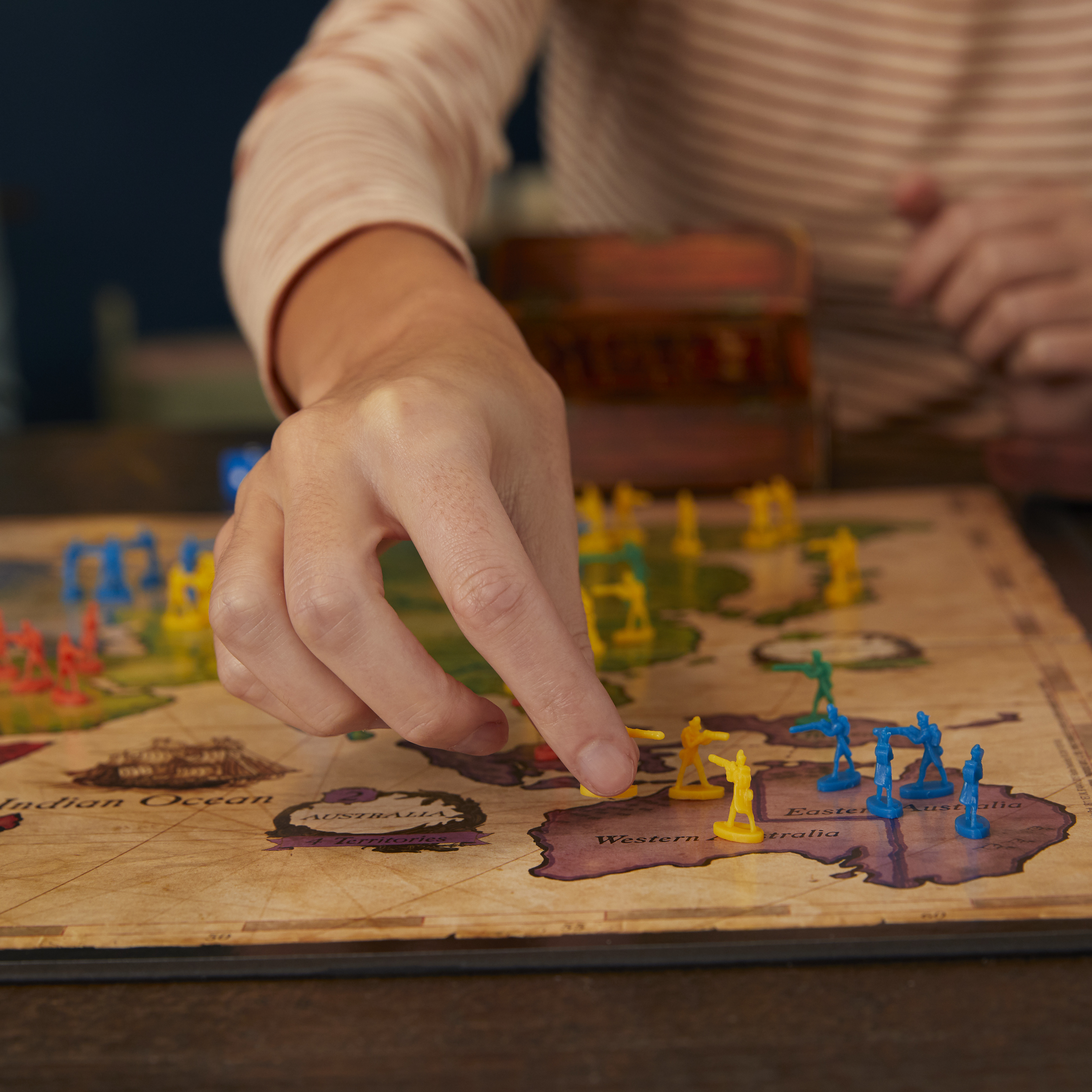Una mano posiziona una figura gialla su una mappa di Risk. Sono visibili altre figure colorate e dettagli della mappa.