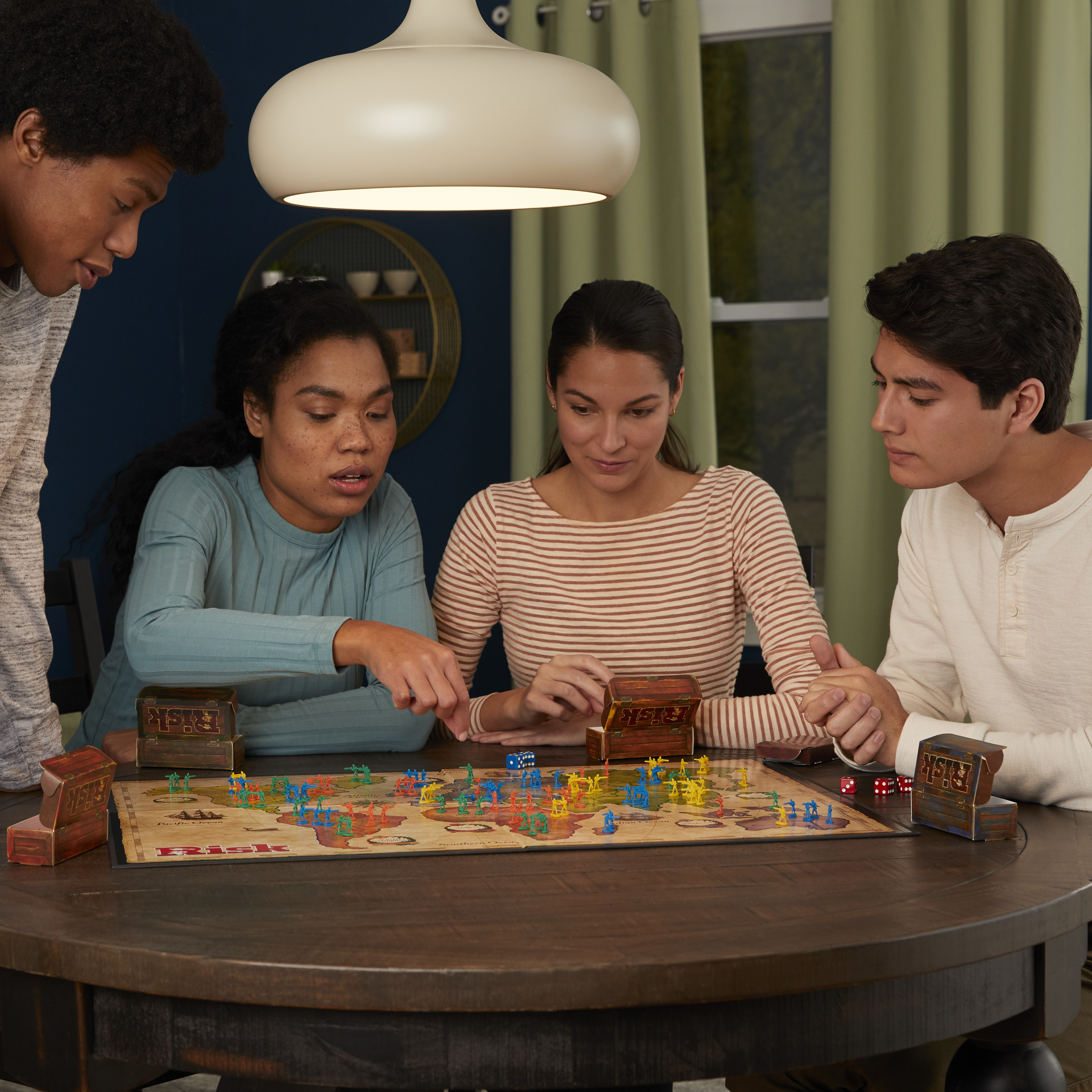Quattro persone giocano a un gioco da tavolo Risk. Dadi e pedine sono sul tabellone. Una luce è appesa sopra.