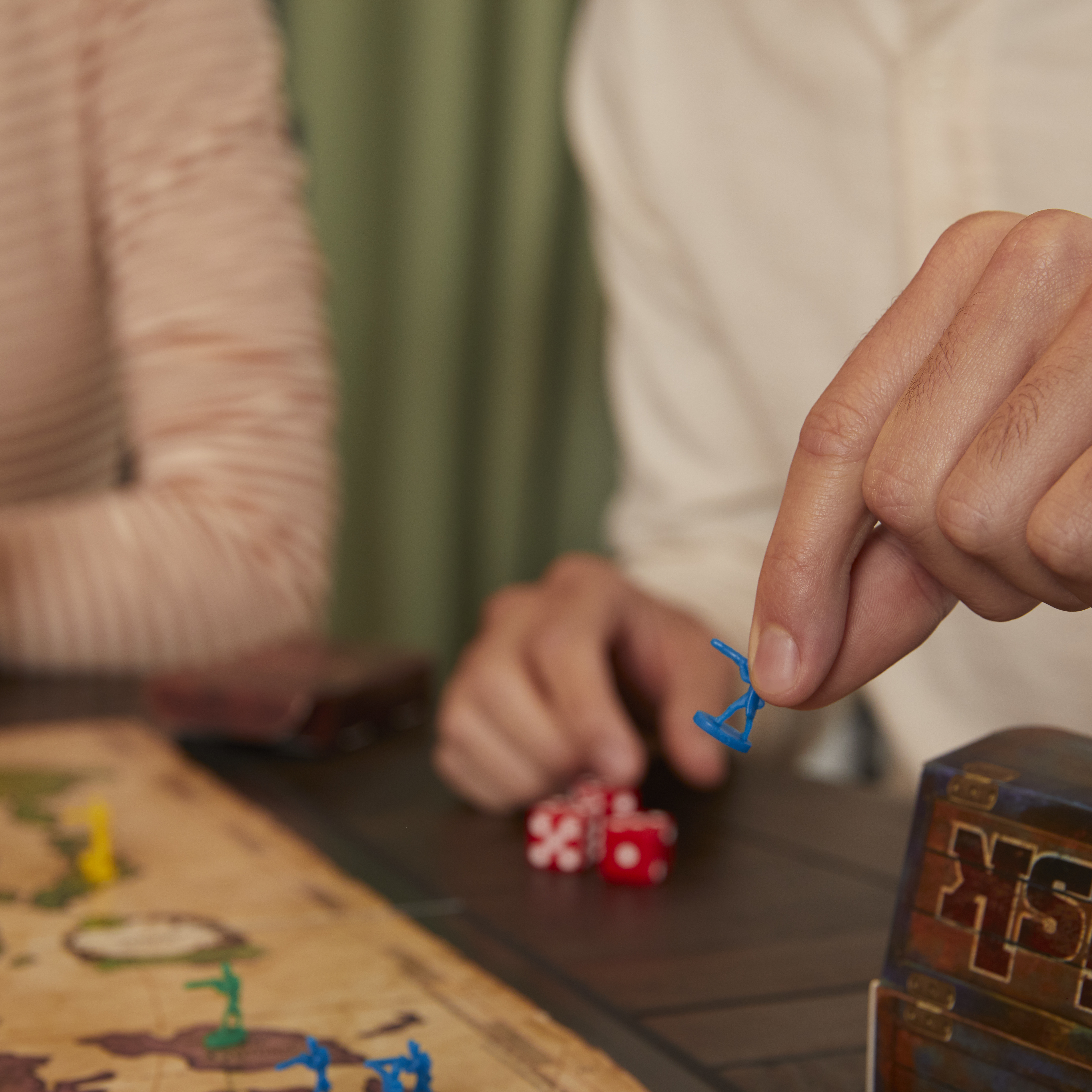 Una persona tiene in mano un pezzo blu del gioco sopra un tabellone di Risk. Dadi e una scatola di gioco sono anche sul tavolo.