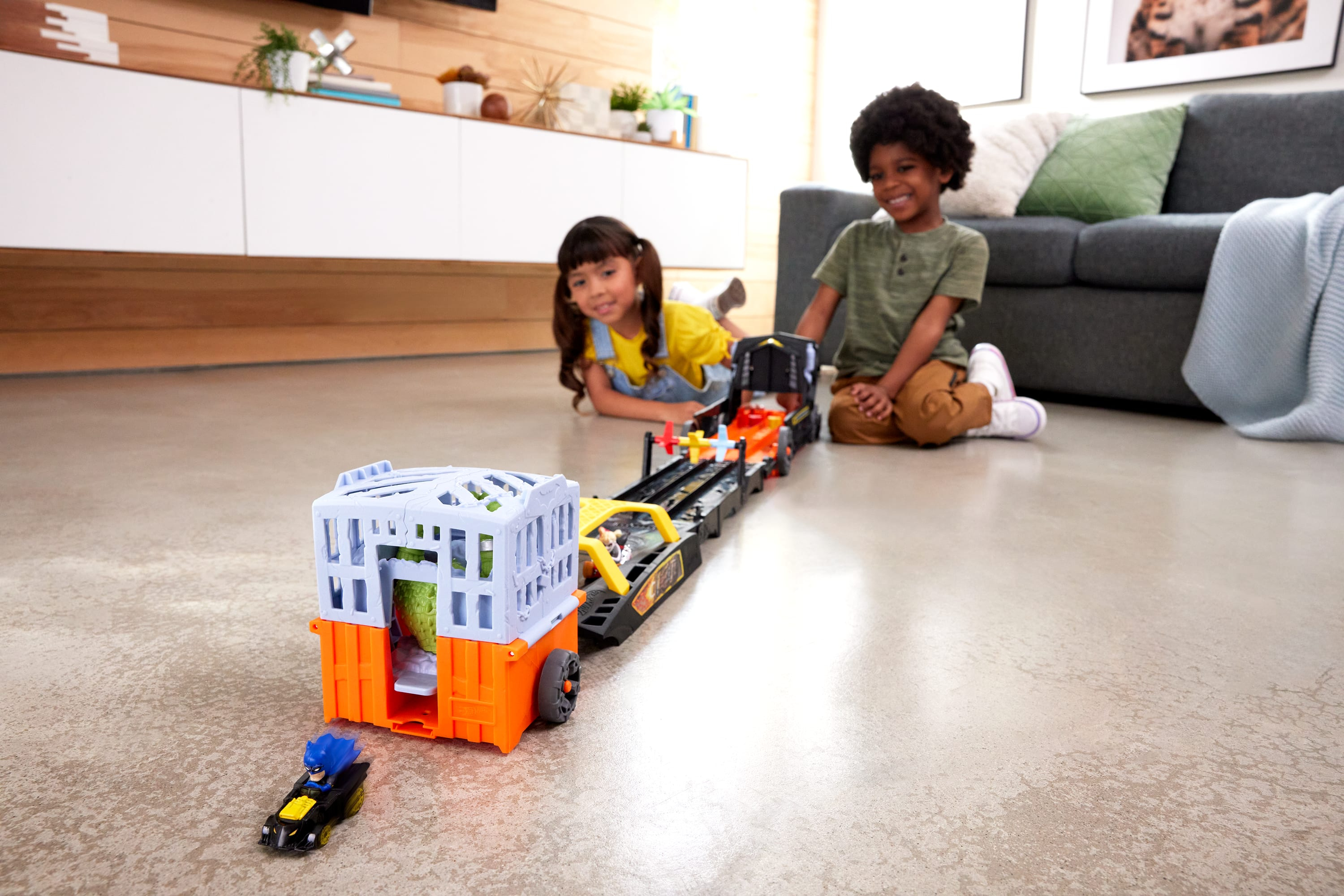 Dwoje dzieci bawi się torem samochodowym. Zabawkowy dinozaur jest w klatce.