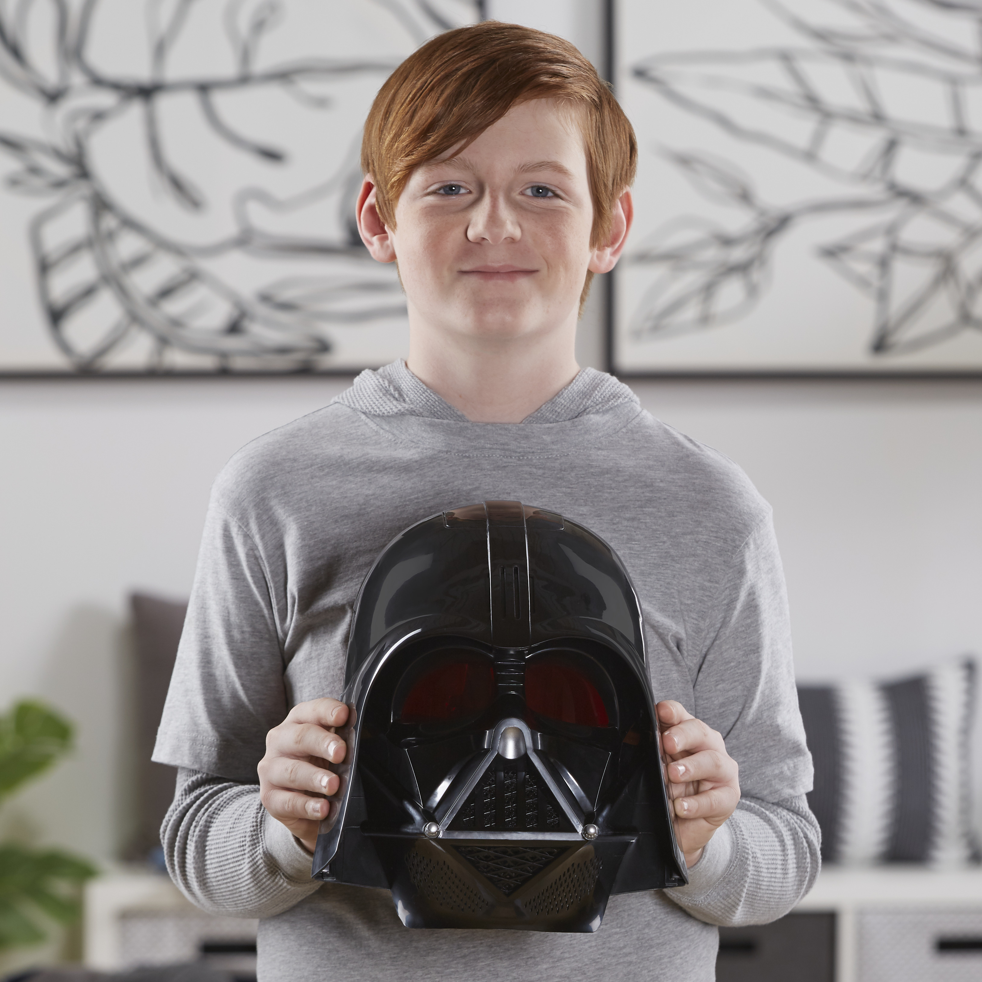 Un niño sostiene una máscara negra de Darth Vader. Está sonriendo. El fondo tiene arte y una estantería.
