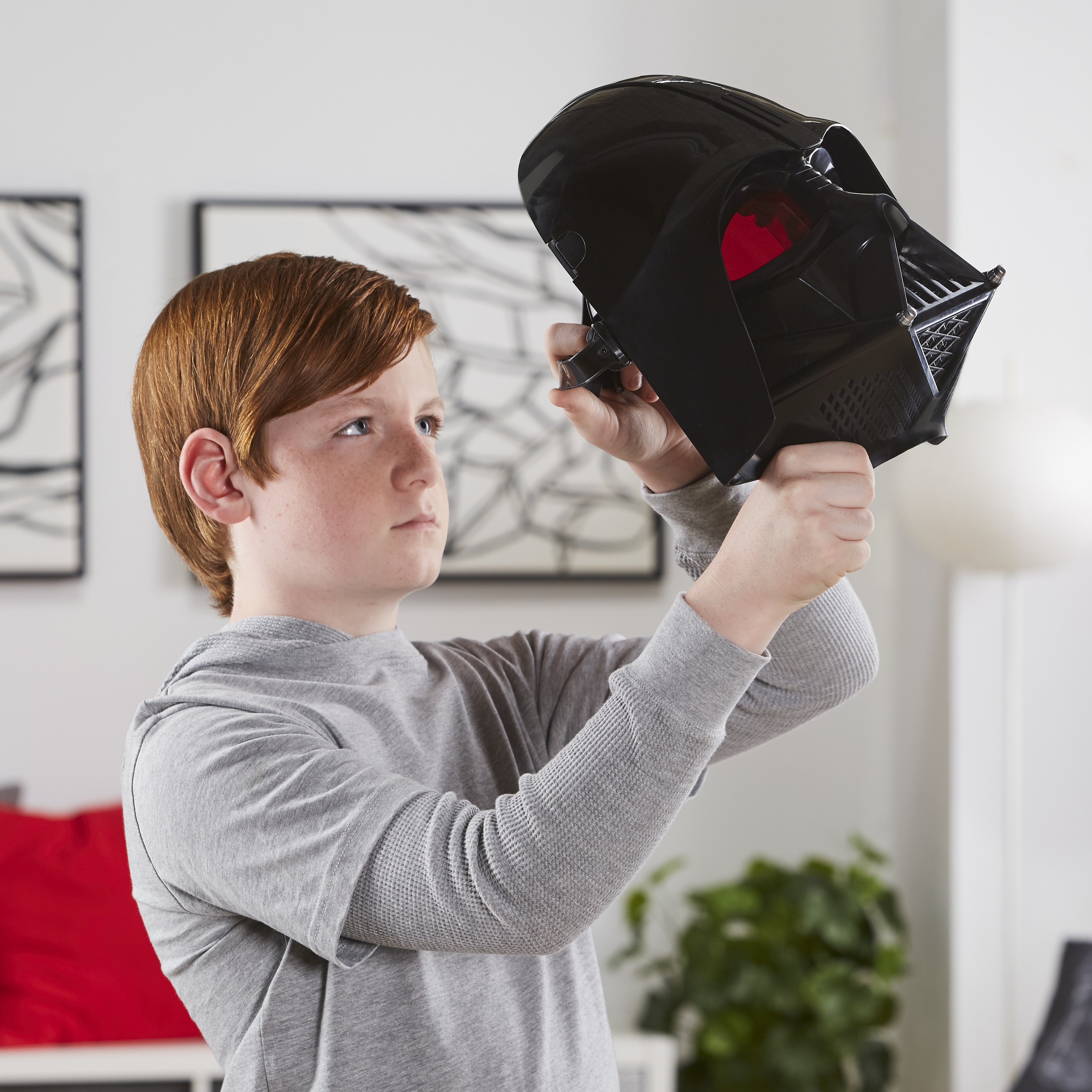 Un niño sostiene una máscara negra de Darth Vader, mirándola. Lleva una camisa gris. El fondo muestra una habitación.