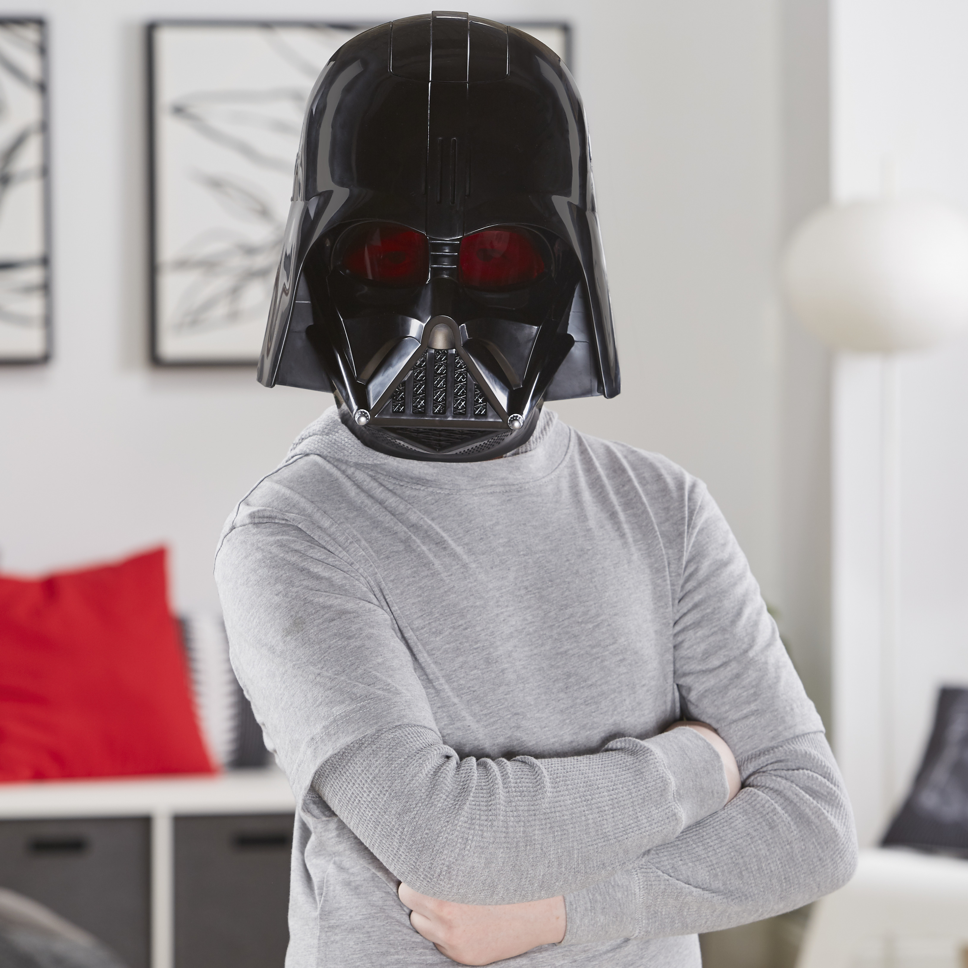 Una persona con máscara de Darth Vader con los brazos cruzados. Camisa gris y fondo con muebles y decoración.