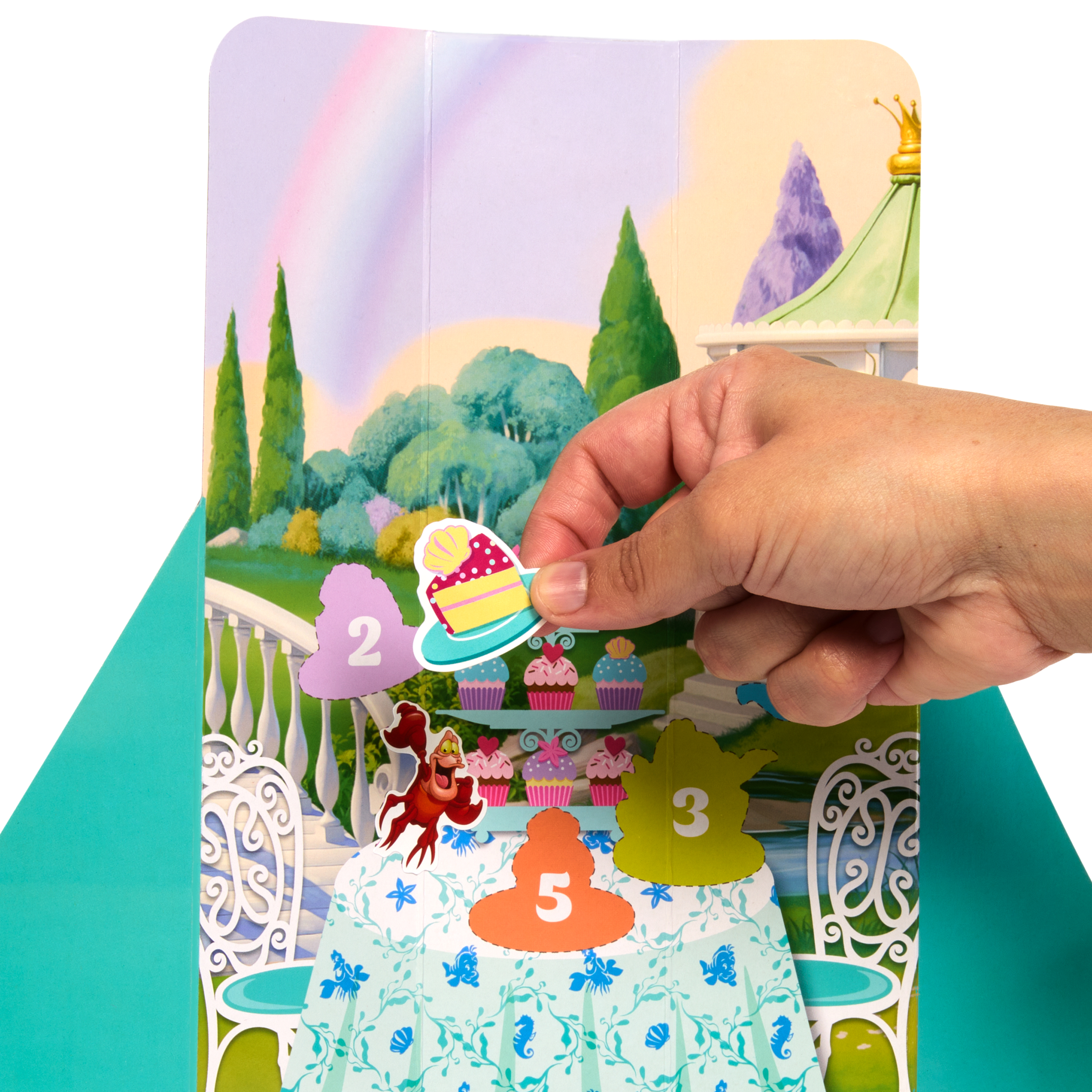 Ręka umieszcza naklejkę z ciastem na karcie ze sceną z bajki, w tym babeczkami i tęczą.