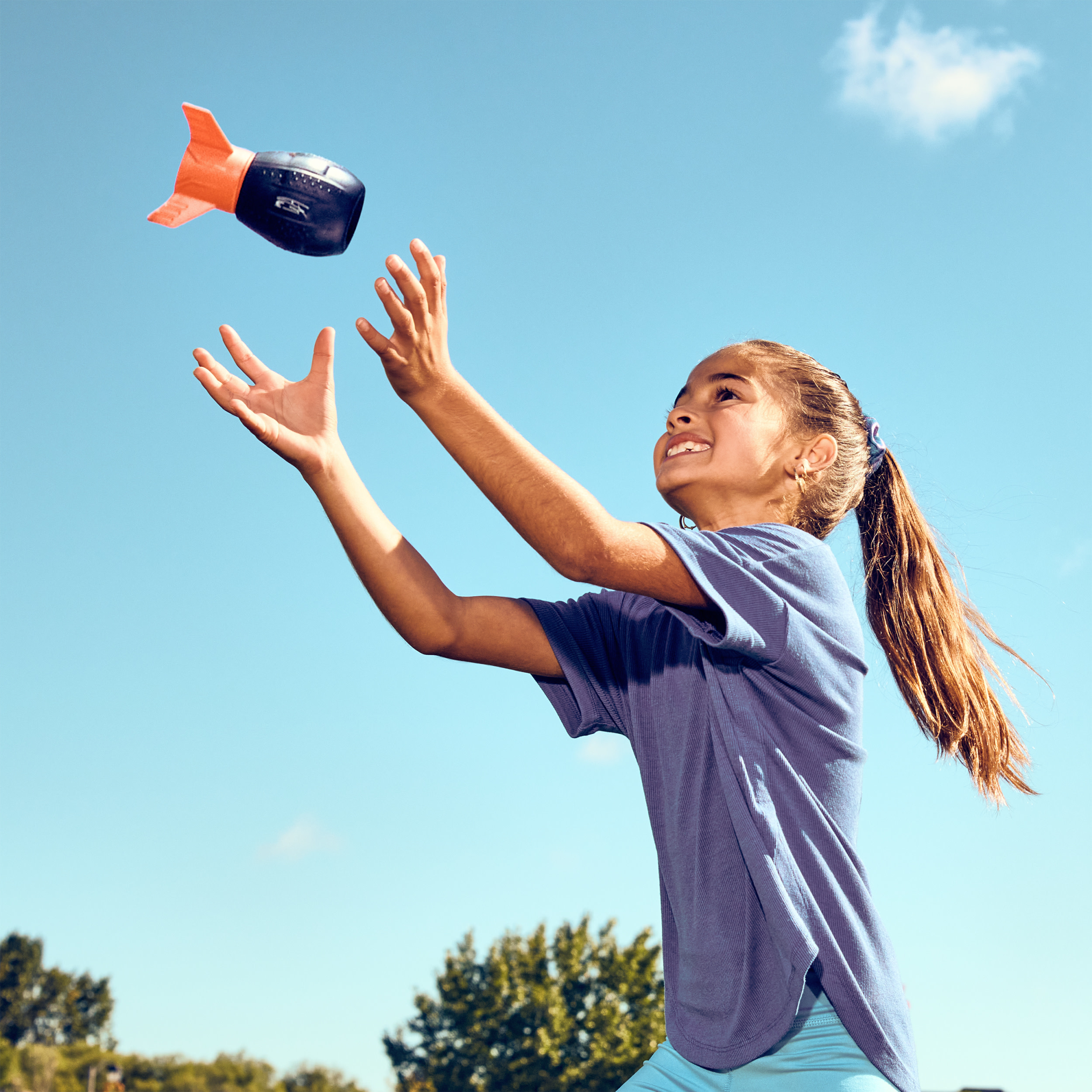 Dziewczyna łapie zabawkę do futbolu na tle błękitnego nieba. Piłka jest czarna i pomarańczowa.