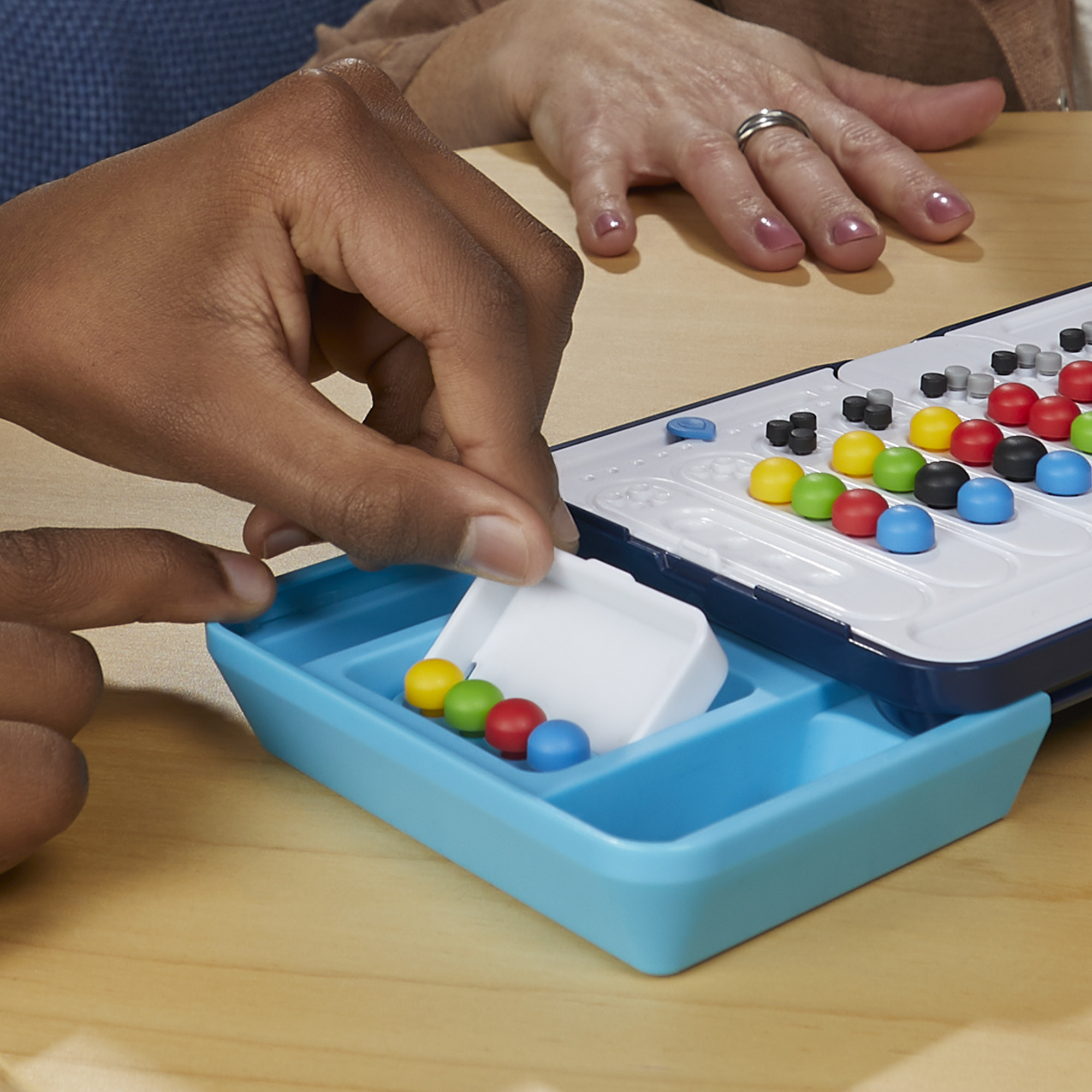 Hände spielen ein Brettspiel. Farbige Stifte und ein Tablett sind sichtbar. Eine andere Hand ist im Hintergrund.
