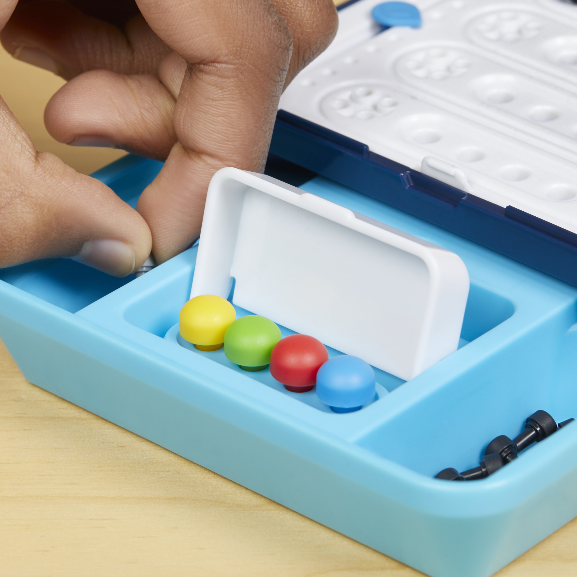 Hand legt ein kleines, graues Teil in ein blaues Spiel. Bunte Stifte sind in einem Fach. Holzoberfläche.