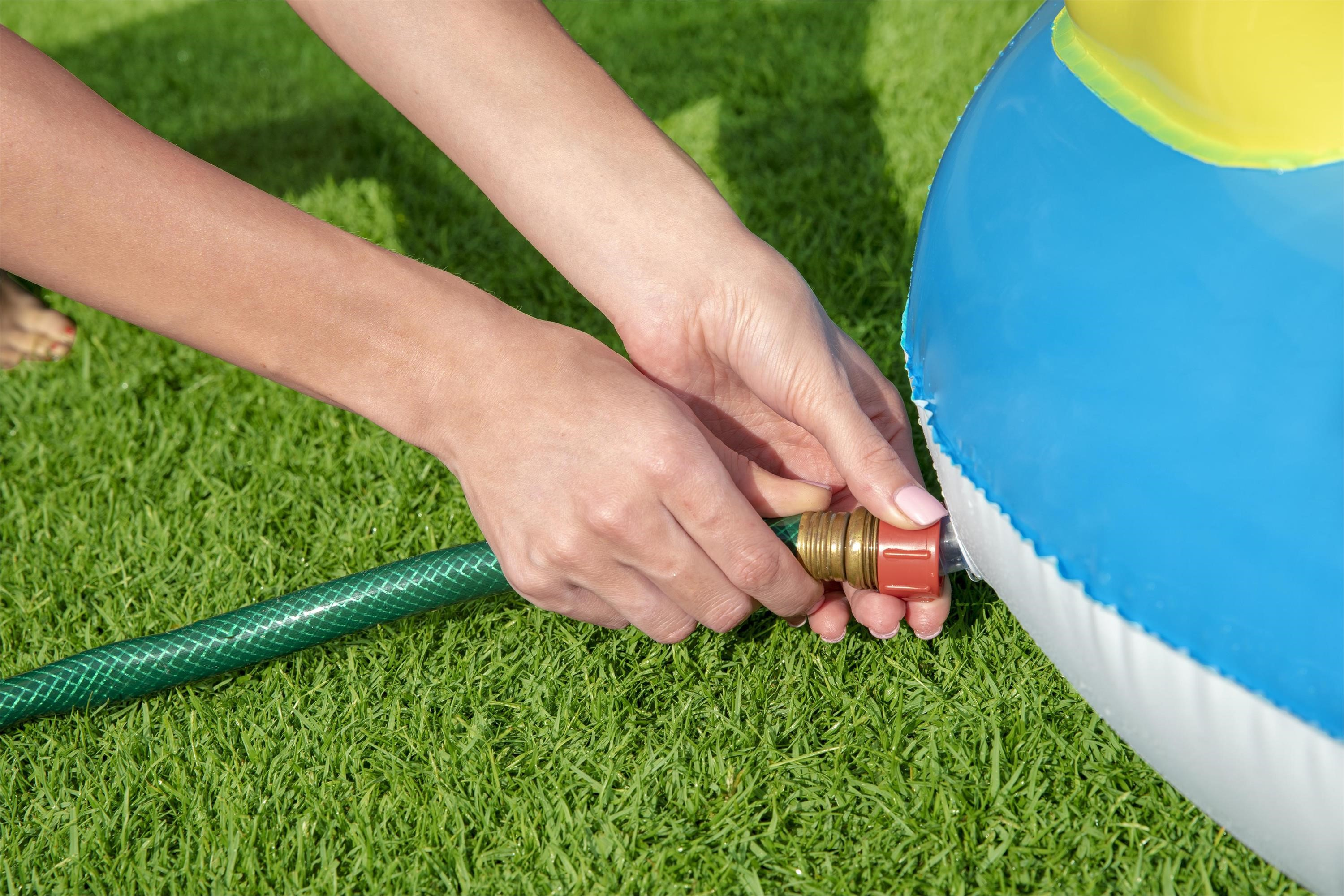 Manos conectan una manguera verde a una piscina inflable azul y amarilla sobre césped verde.