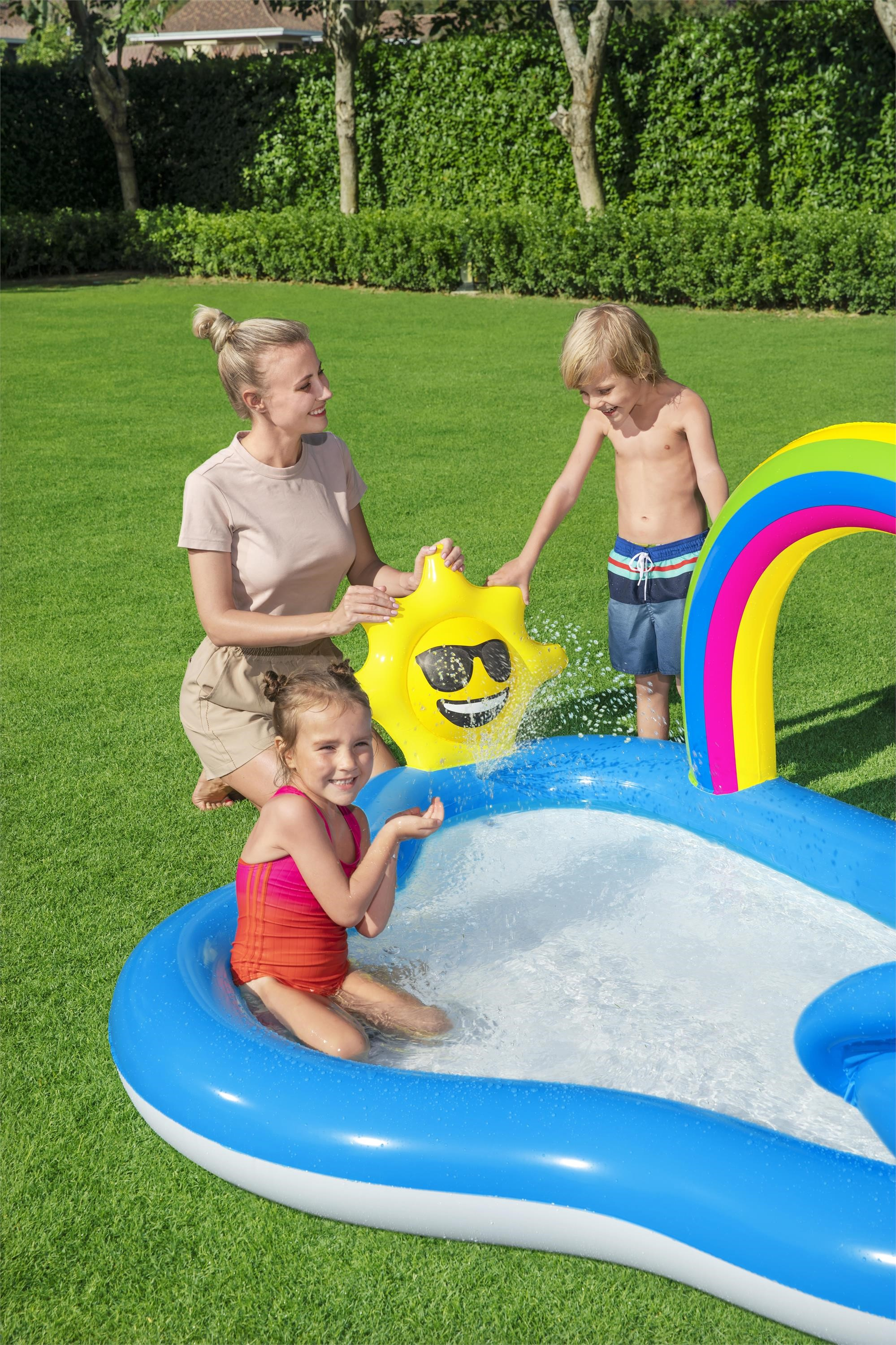 Una mujer y dos niños juegan con un aspersor en forma de sol en una piscina inflable azul sobre césped.