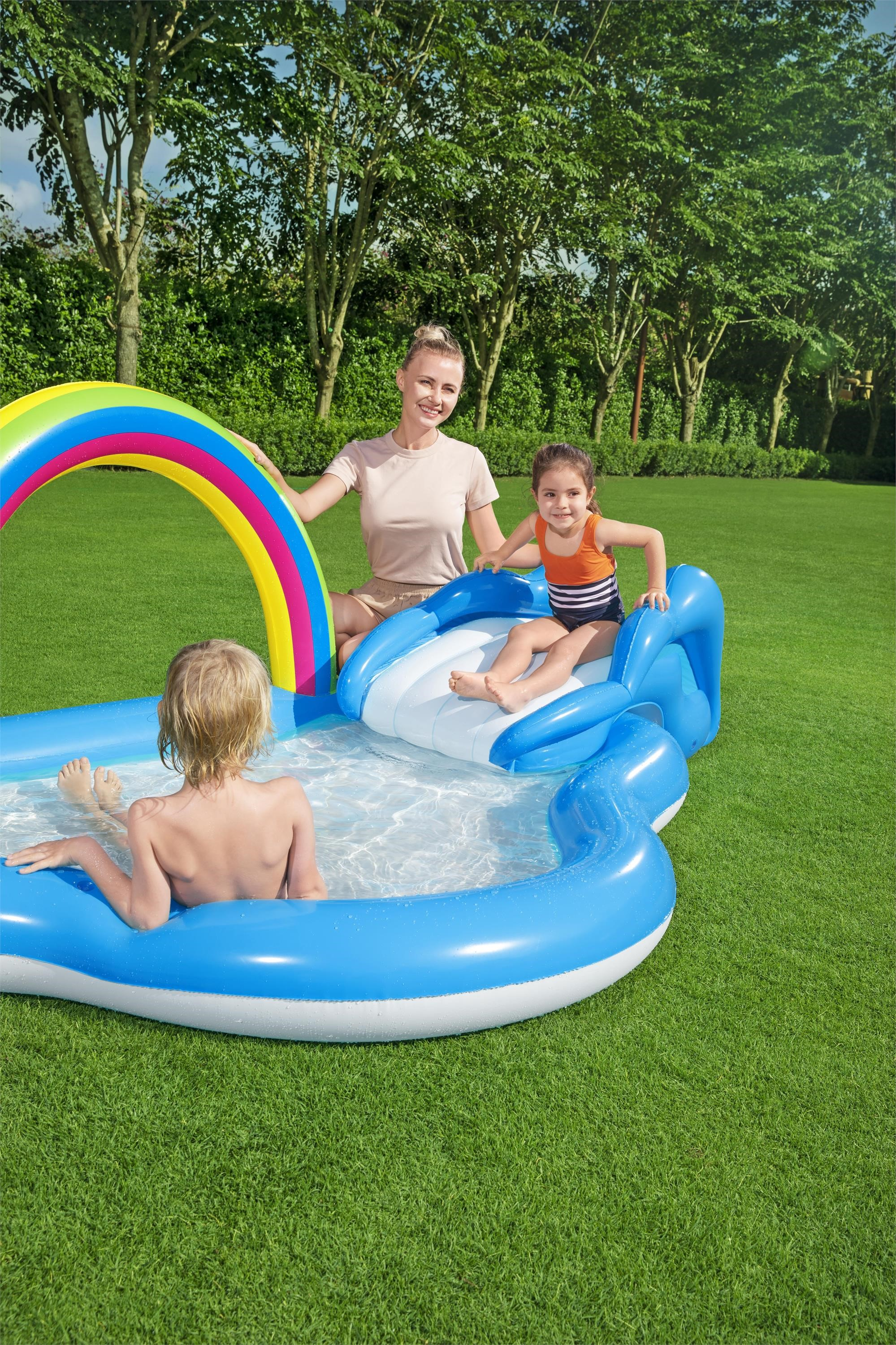 Una mujer y dos niños juegan en una piscina inflable azul con un arco iris en un césped verde.