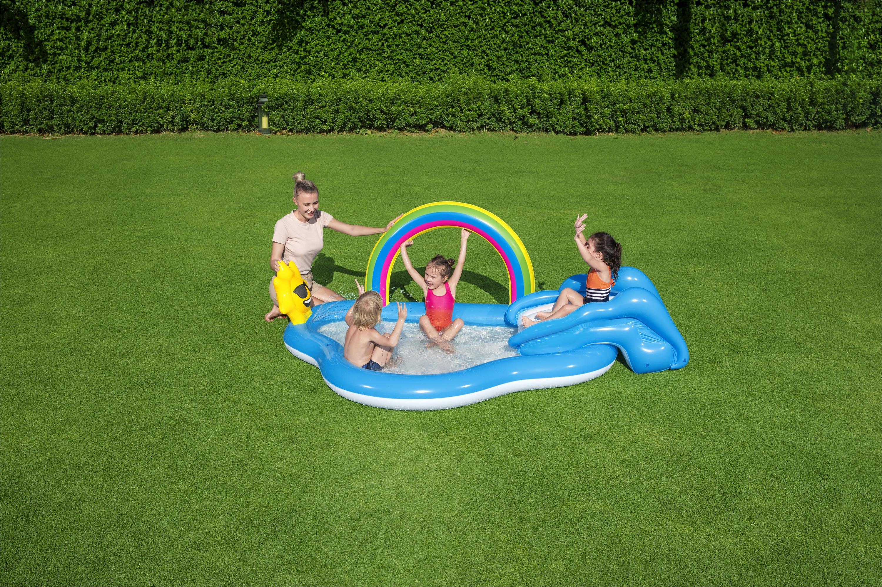 Una mujer y tres niños juegan en una piscina inflable azul sobre un césped verde. Un seto en el fondo.