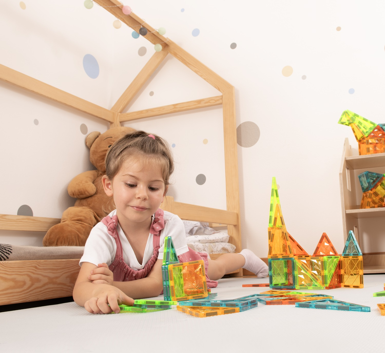 Una bambina gioca con blocchi colorati sul pavimento. Un orsacchiotto e una cornice di casa in legno sono dietro.