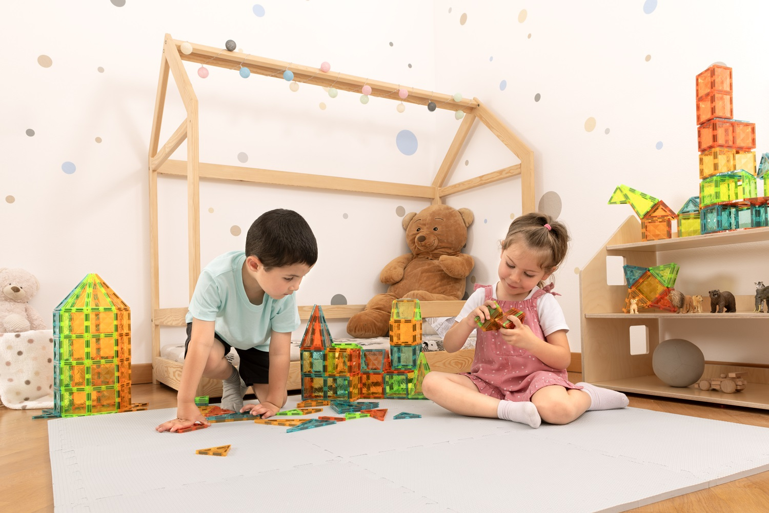 Due bambini giocano con blocchi magnetici colorati in una stanza. Un orsacchiotto è dietro di loro.