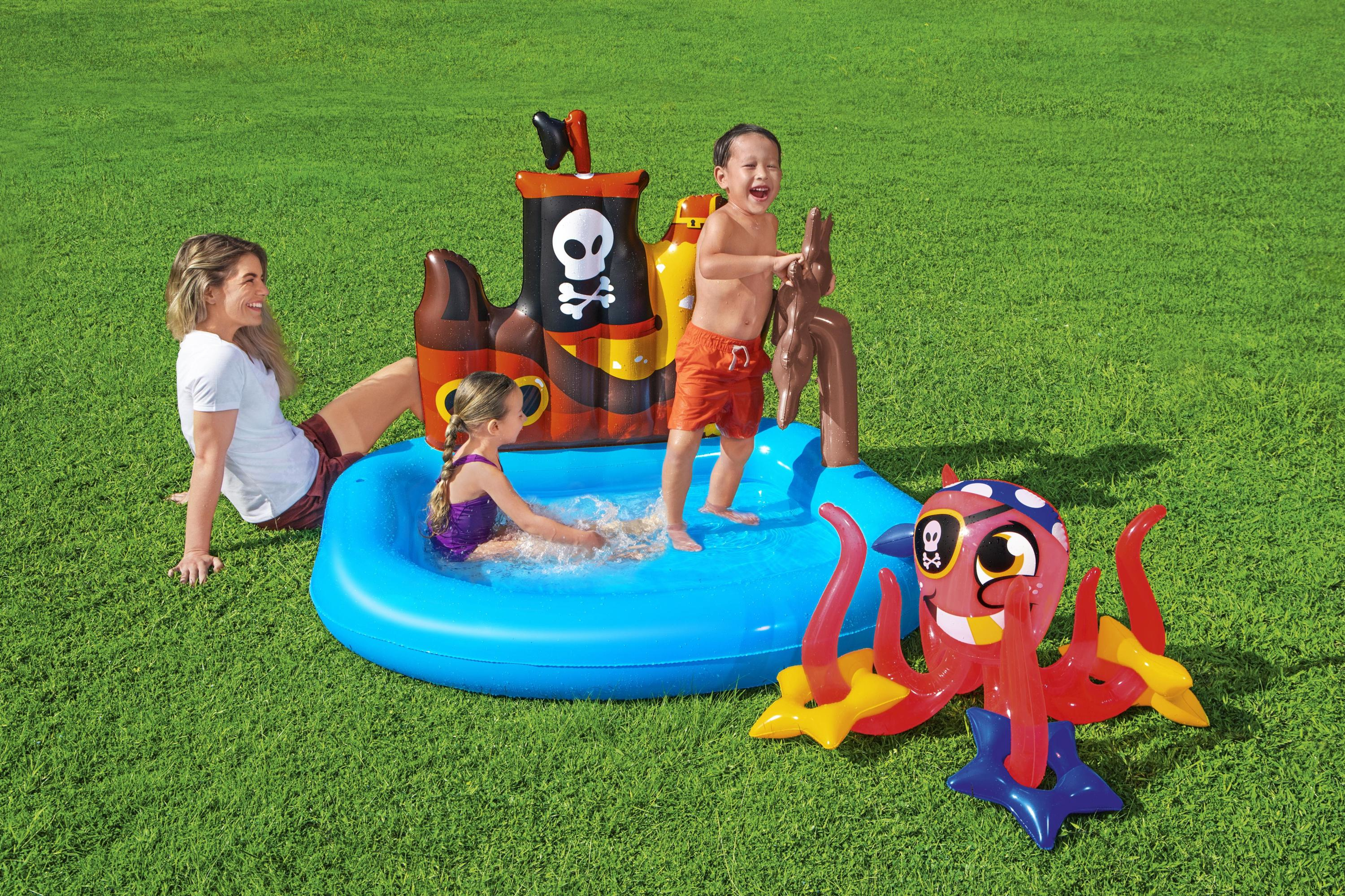 Kinder spielen in einem Piratenschiff-Pool auf Gras. Eine Frau schaut zu. Ein aufblasbarer Oktopus ist in der Nähe.